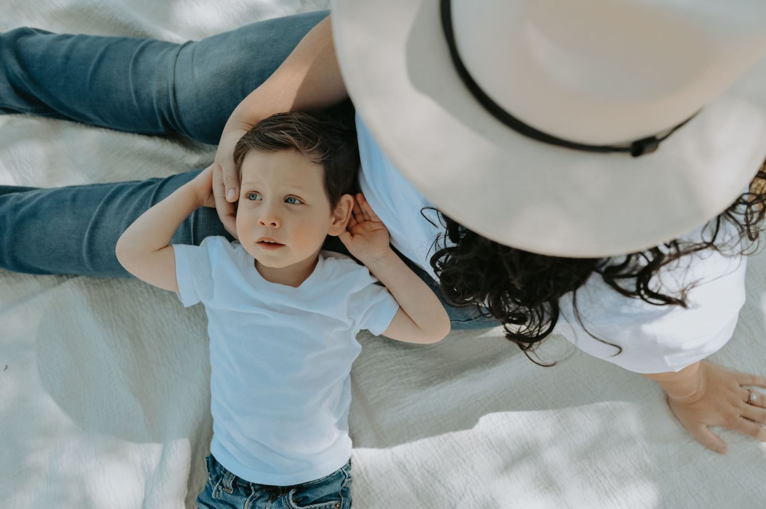 A young toddler boy in a white shirt lies on a blanket beside a woman in a white hat outdoors.