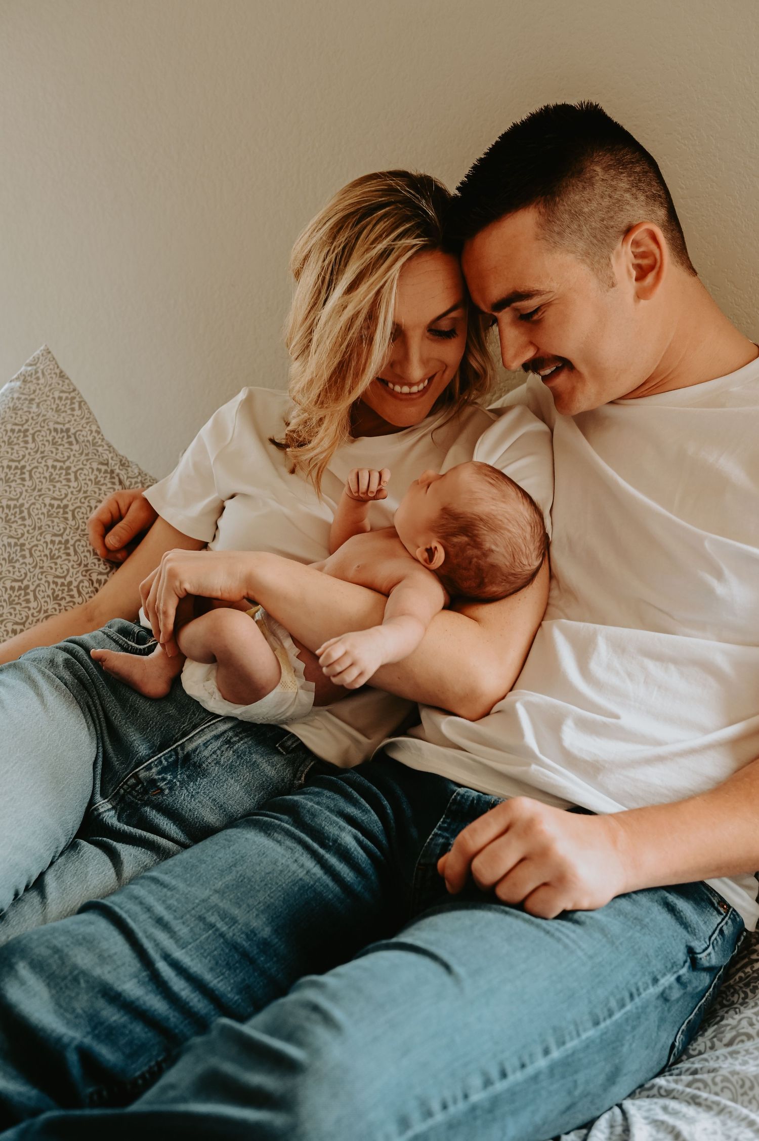 Happy couple in white tees and jeans cuddling newborn baby on a cozy bed at home.