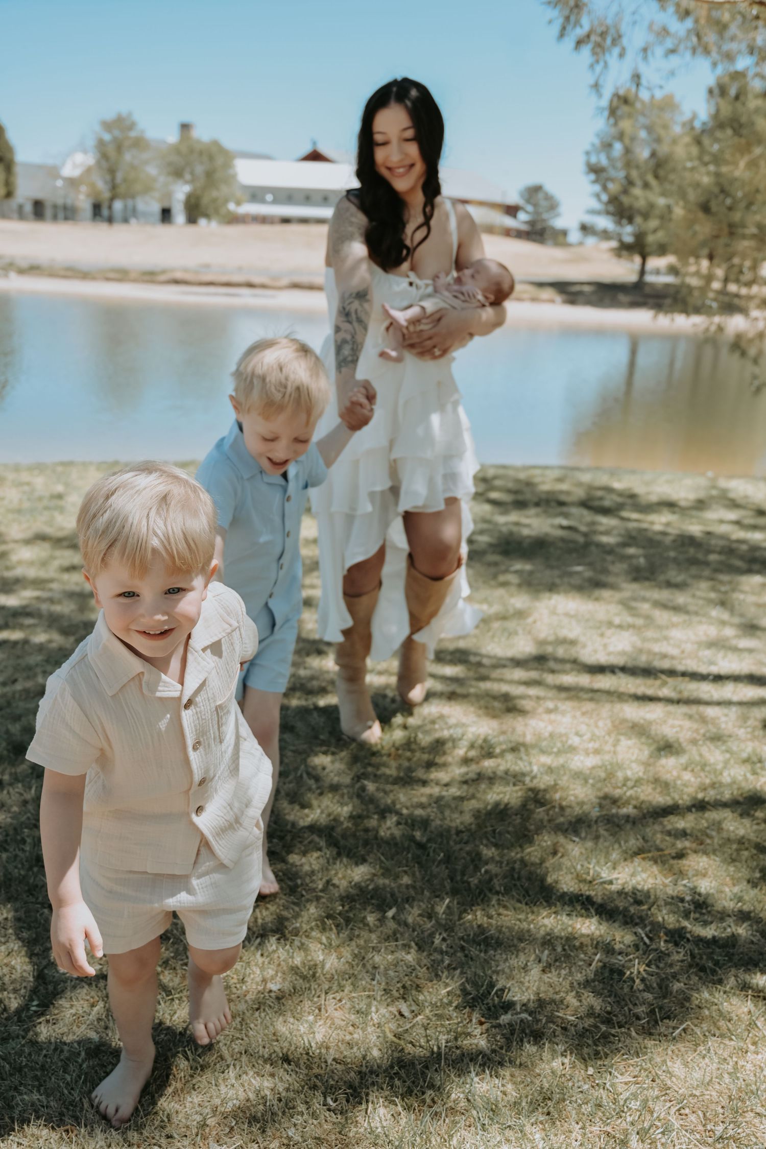 A mother in a white dress holds a newborn outdoors near a pond while two young boys walk ahead on grass.