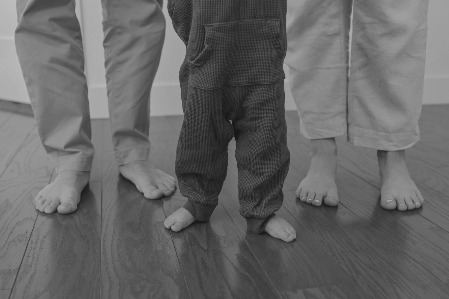 Black and white photo of two adults and a child standing barefoot on hardwood floor, showing only their legs and feet.