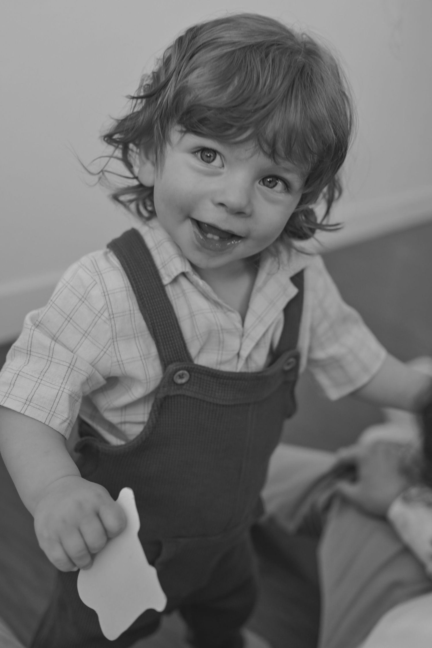 Smiling toddler in overalls and plaid shirt holding a toy in a black and white portrait photo.