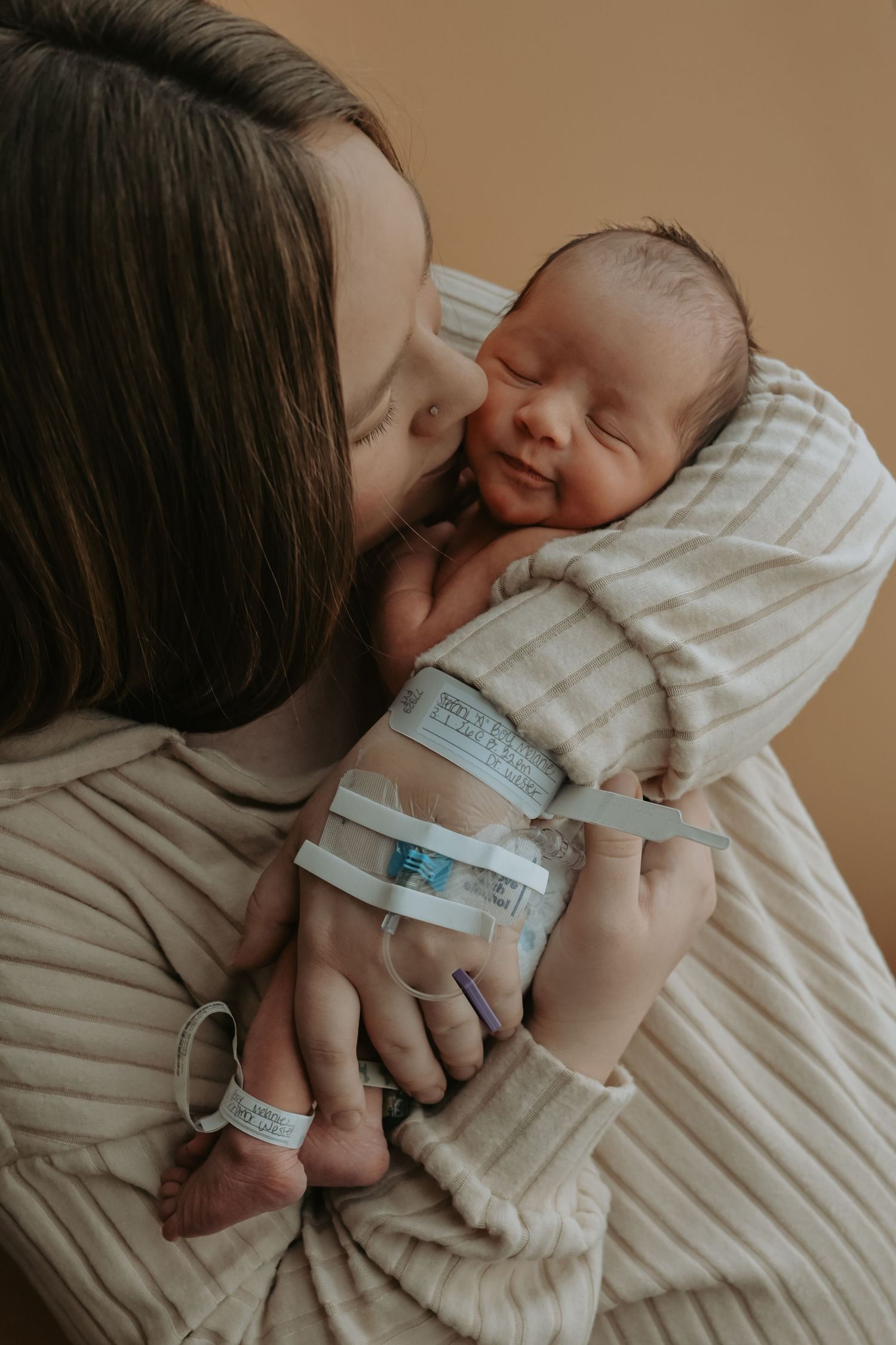 Mother tenderly kissing her sleeping newborn baby wrapped in a striped blanket with hospital ID bands.