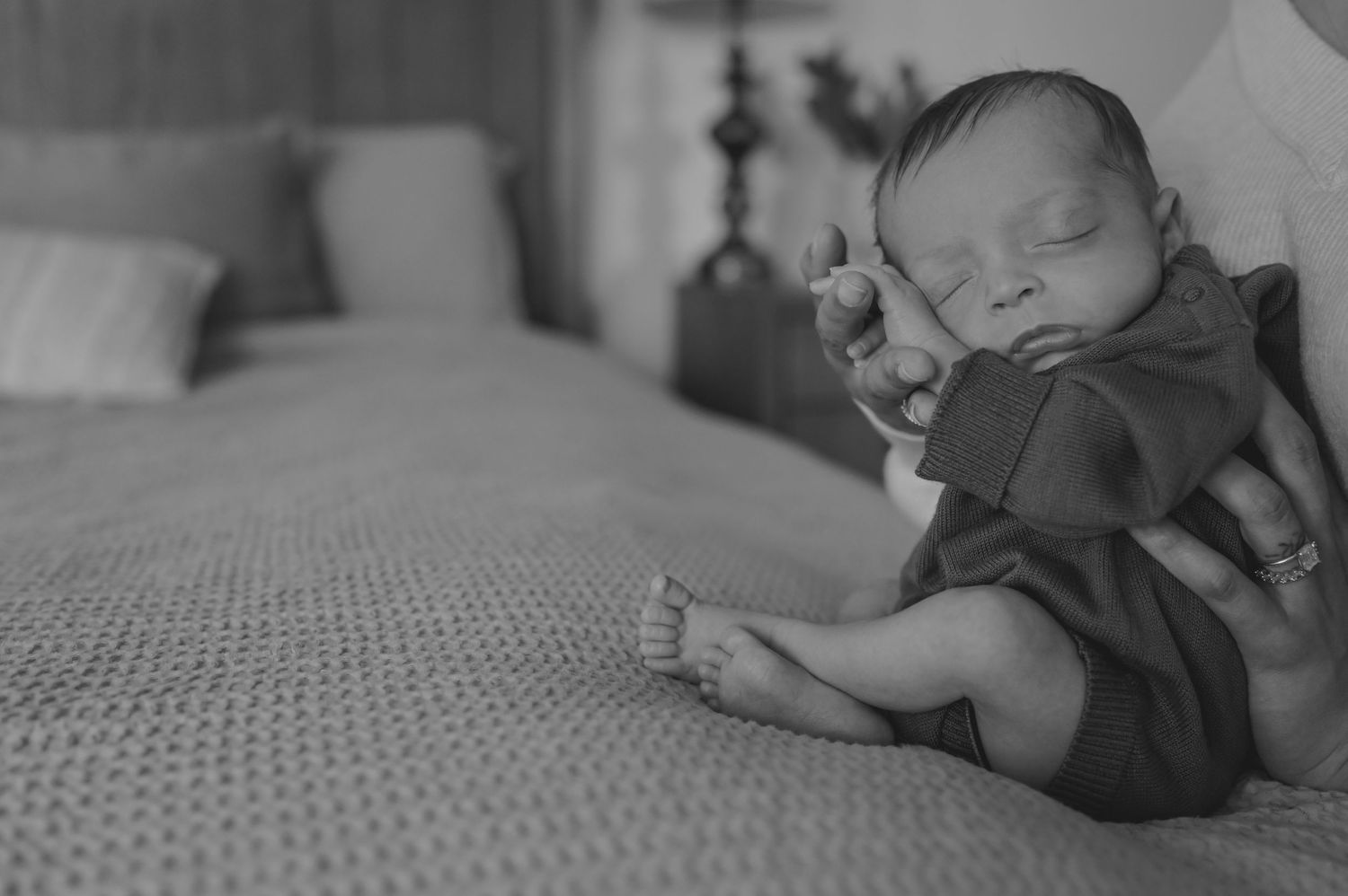 Black and white photo of a sleeping newborn baby in a dark romper, curled up on a textured blanket near a bed.