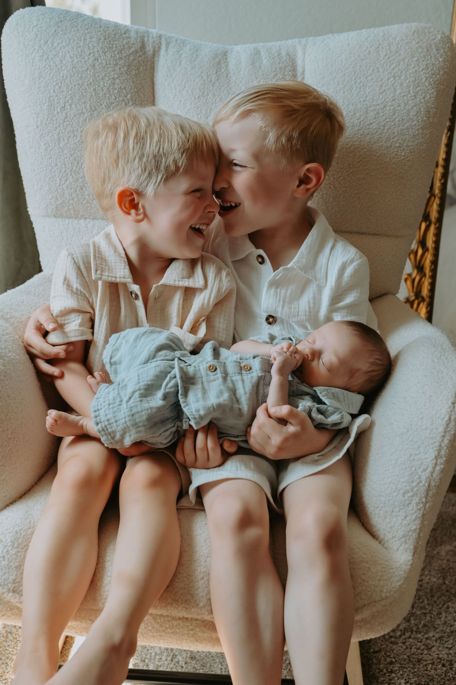 Two young blonde boys sit in a white chair, smiling at each other while holding a newborn baby sibling.