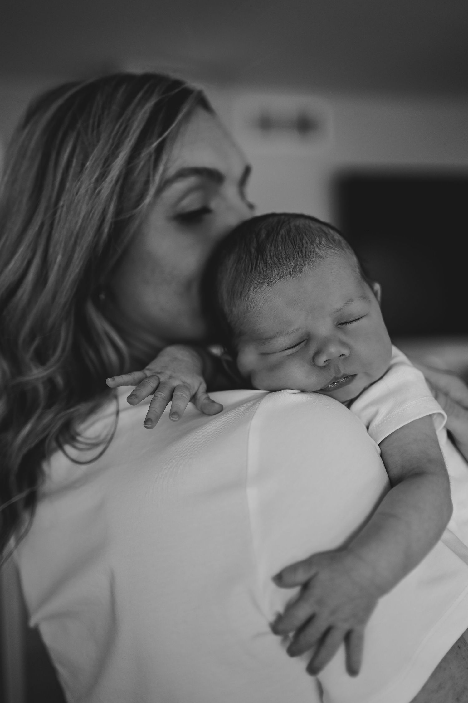 Black and white photo of a mother tenderly kissing her sleeping newborn baby wrapped in a white blanket.