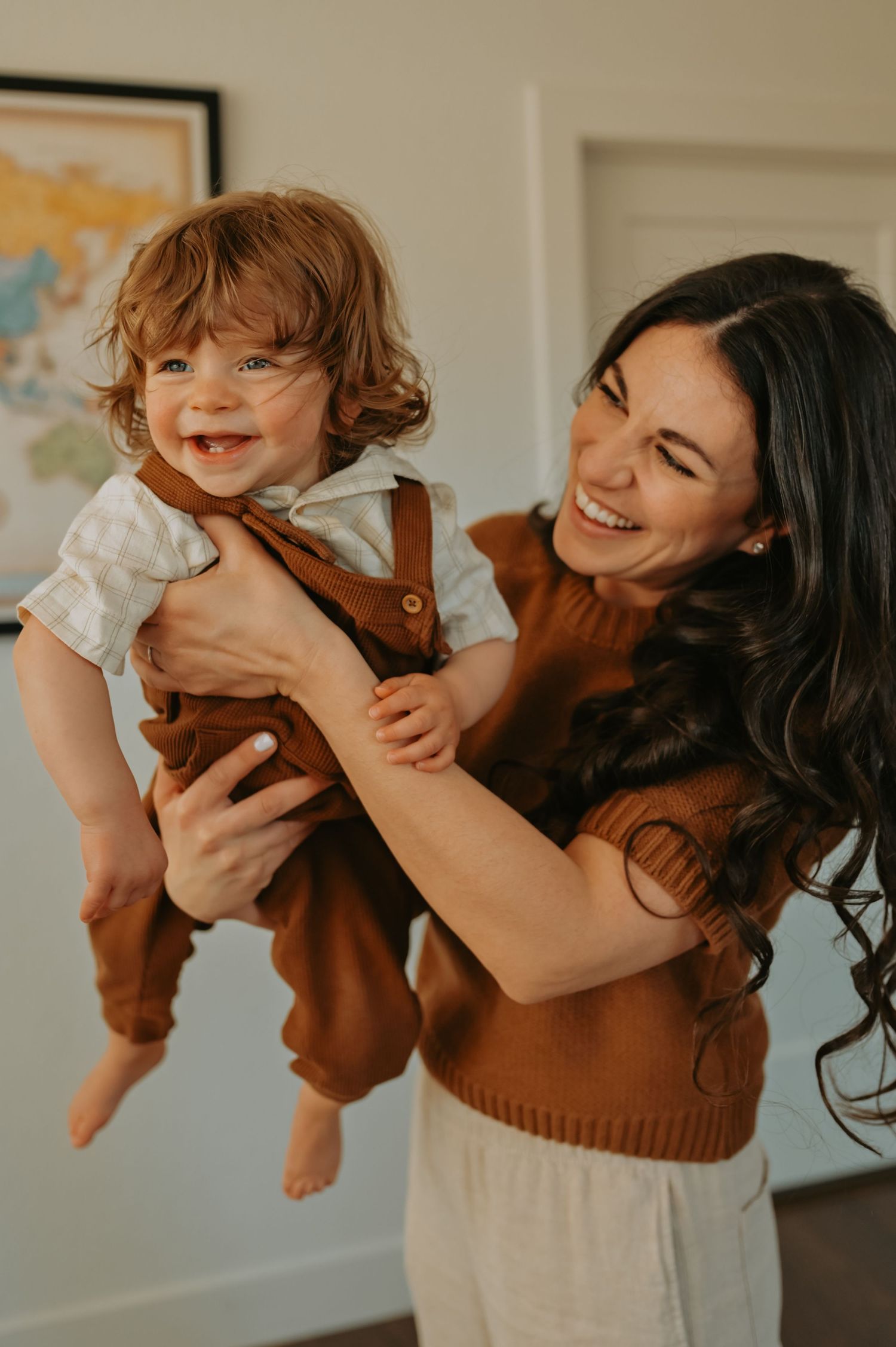 A laughing mother lifts her smiling toddler in brown overalls indoors, both sharing a joyful moment together.