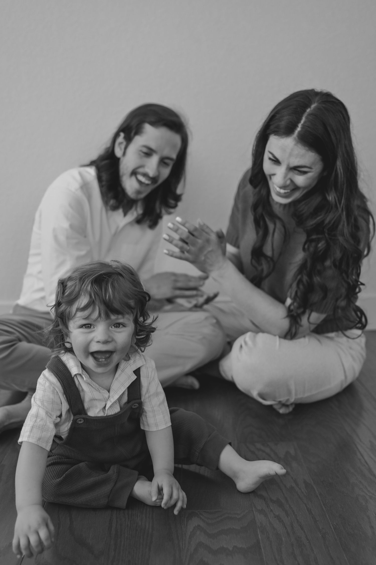 Black and white photo of a happy young family with a laughing toddler sitting on a hardwood floor together.