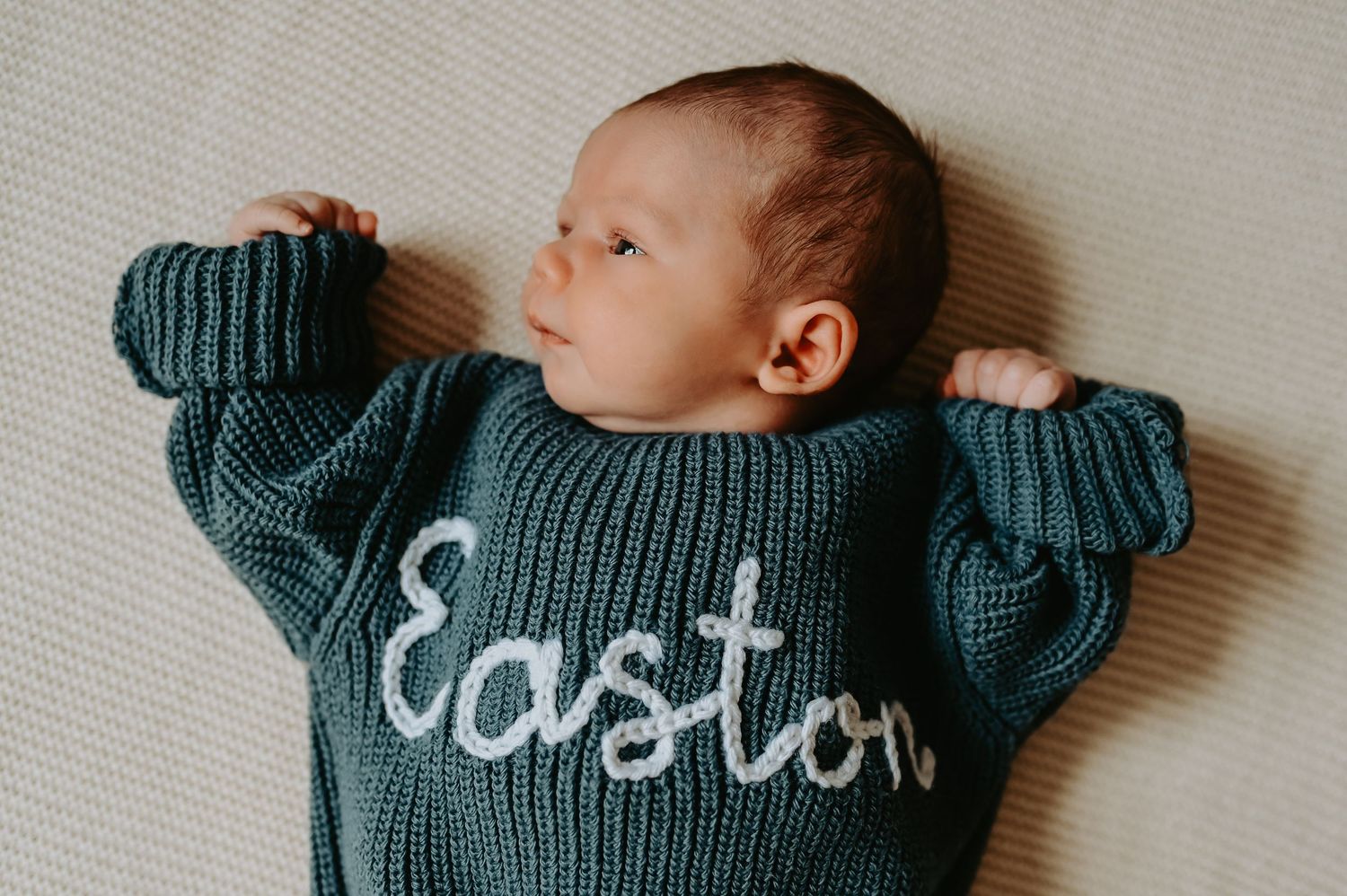 Newborn baby named Easton lying on back wearing a teal knit sweater with name embroidered in white script.