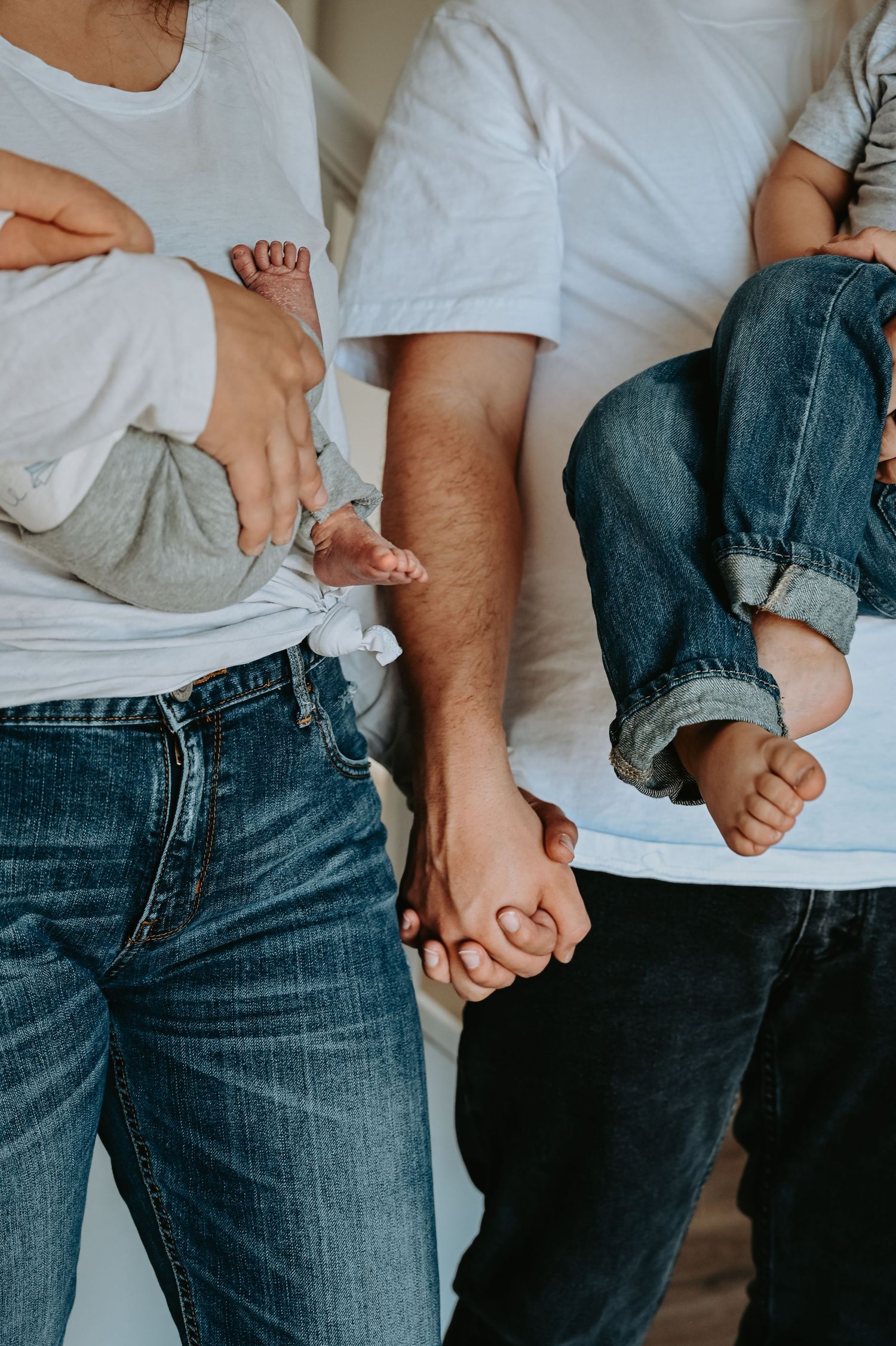 Two parents in white tees and jeans holding their newborn and toddler children, showing family hands and tiny feet.