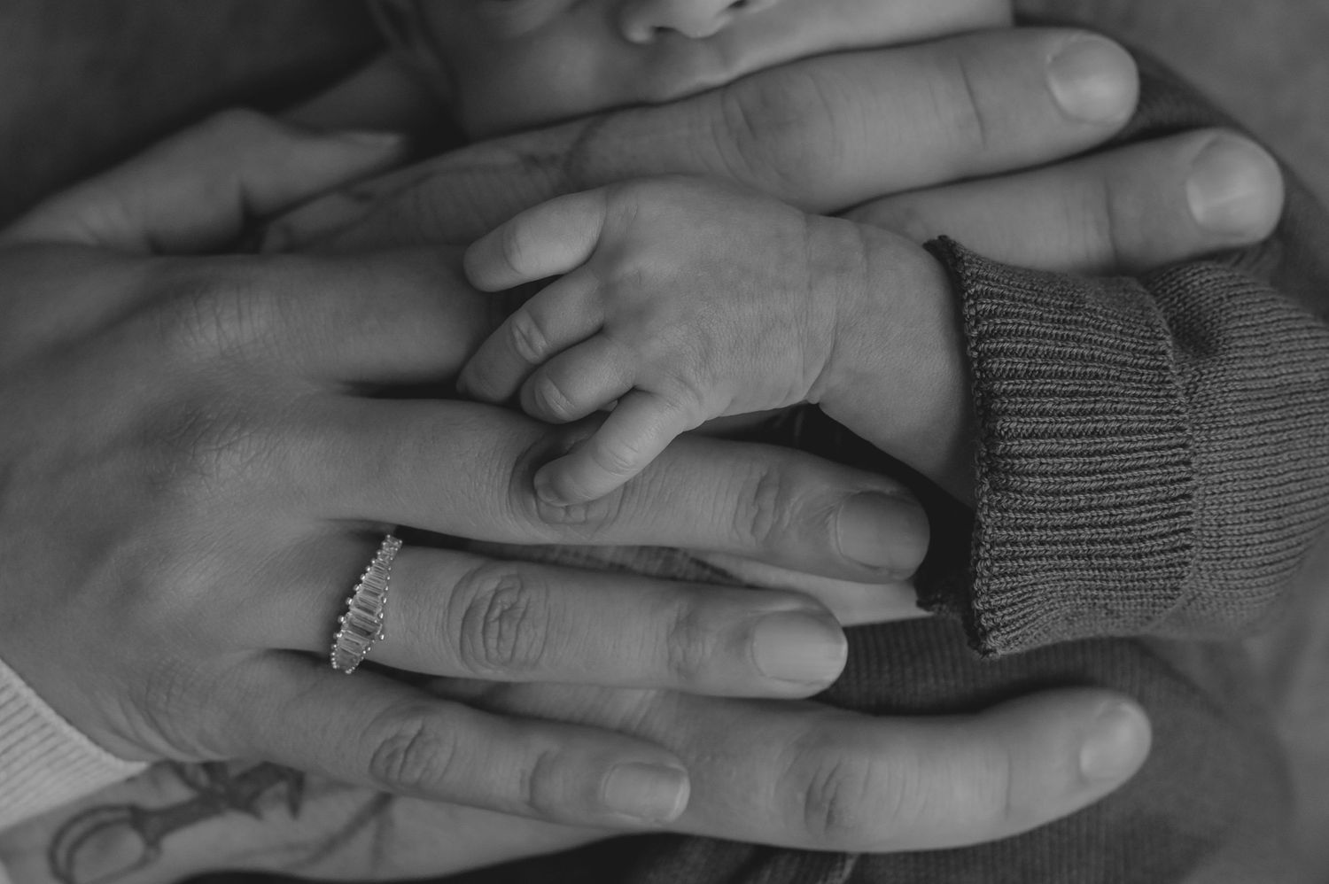Black and white photo of newborn baby hand resting on parents hands, one wearing a ring and tattoo.