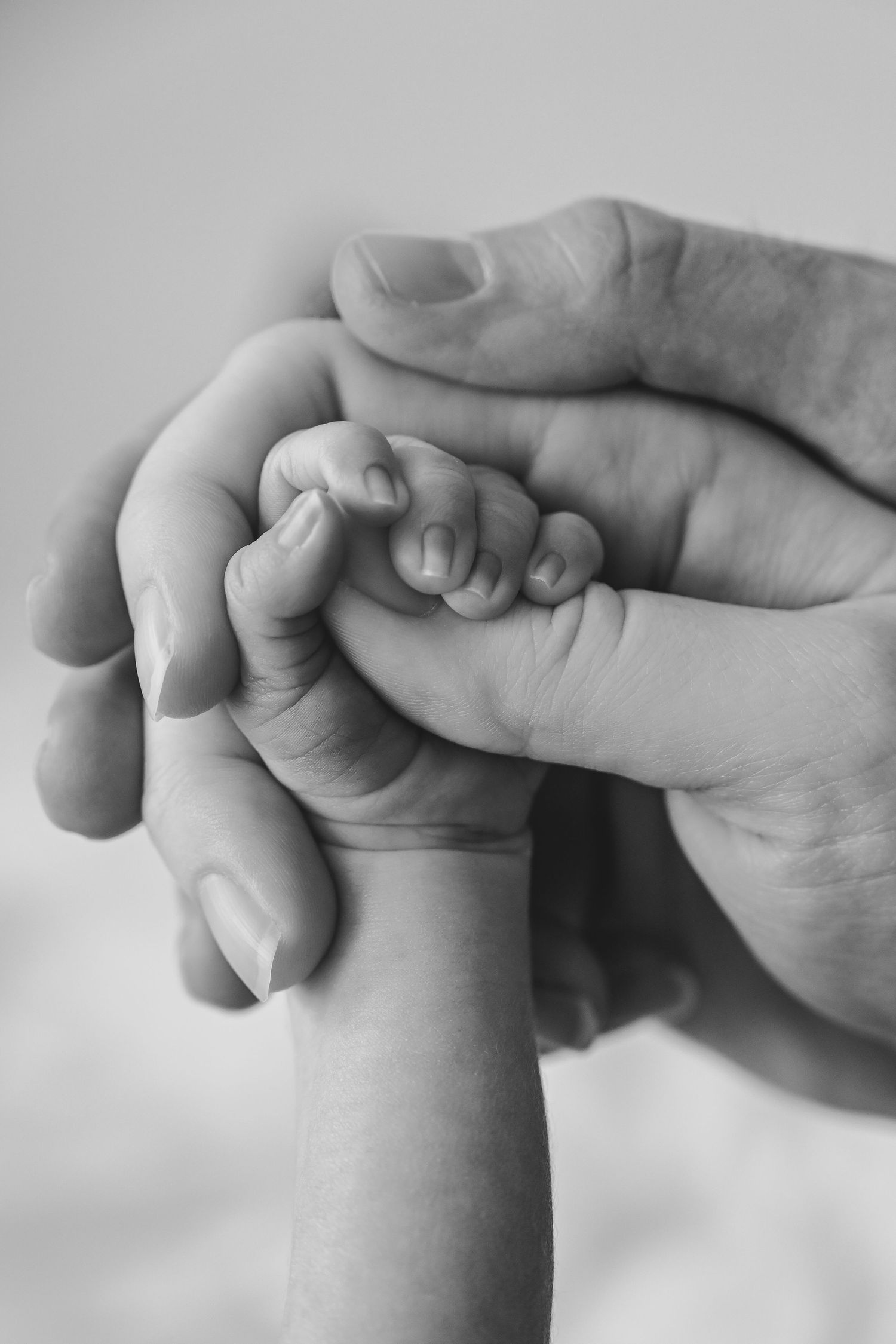 Black and white close-up of a newborn baby's tiny hand gently grasped by two adult hands.