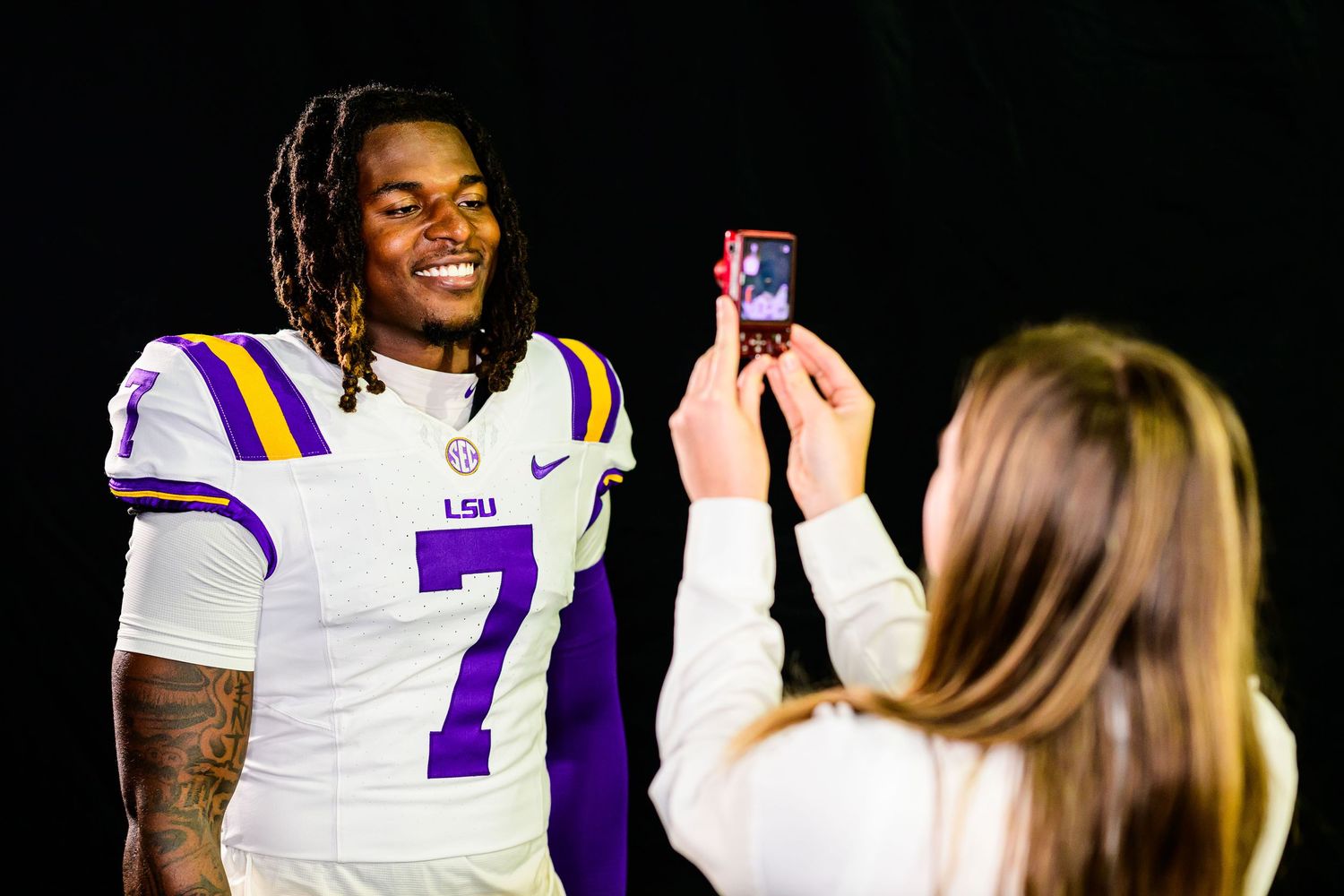 LSU football player wearing number 7 white jersey poses while a woman photographs him with a smartphone.