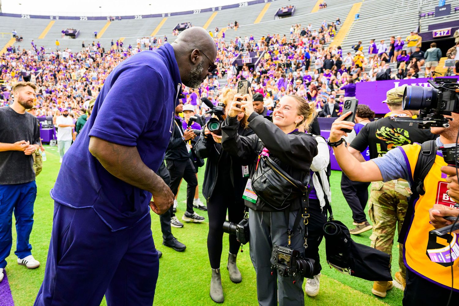 Tall man in purple shirt greets photographer on football field with purple-clad fans in stadium stands.