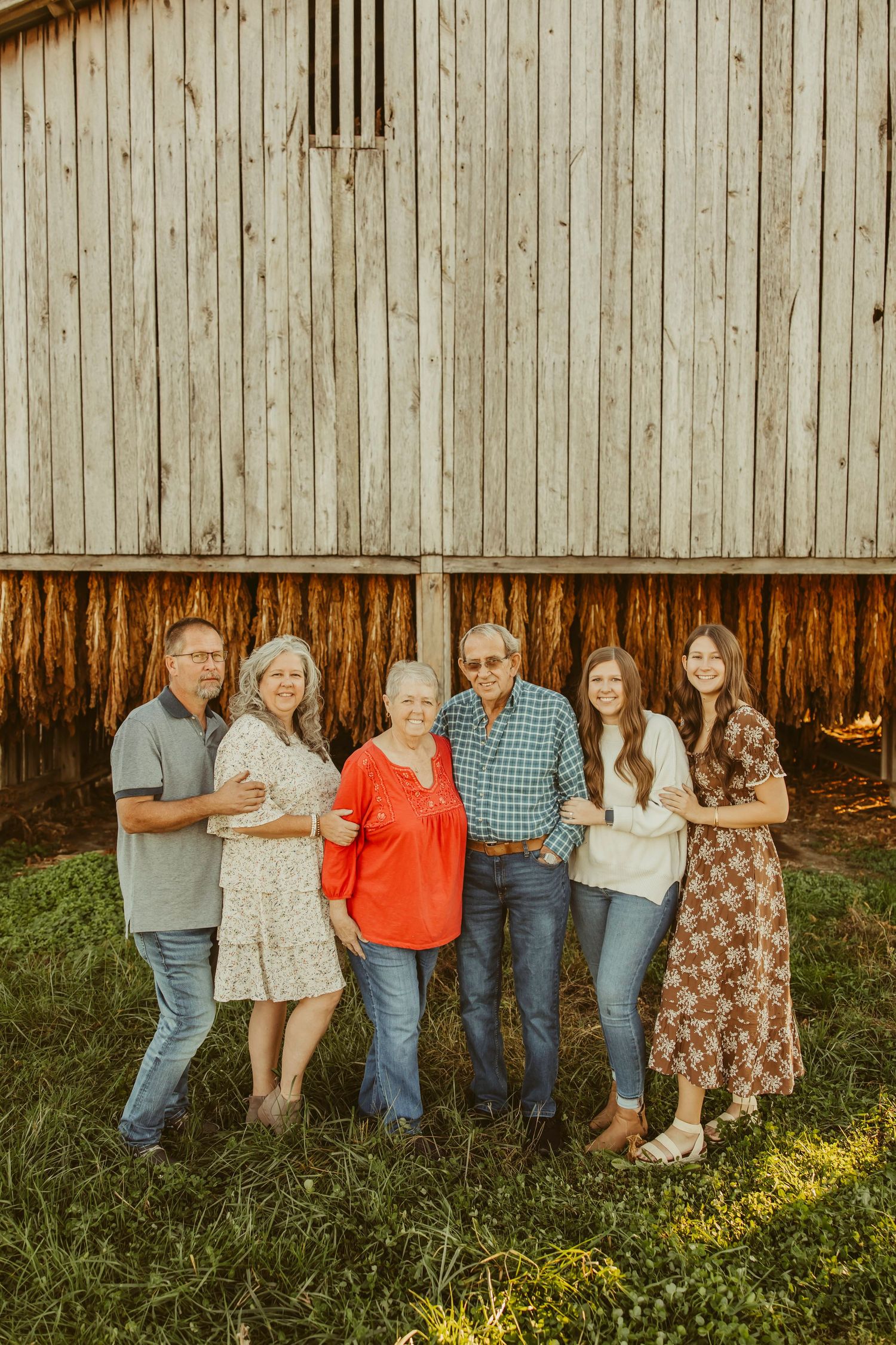 Six family members smiling together outdoors in front of a rustic wooden barn on a sunny day.