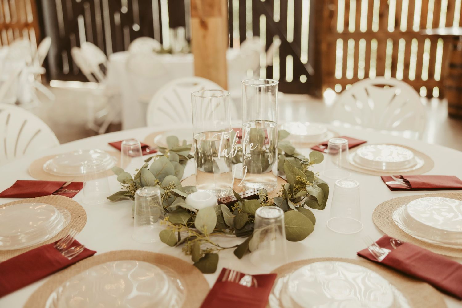 Elegant wedding reception table with eucalyptus wreath centerpiece, candles, white plates, and crimson napkins.