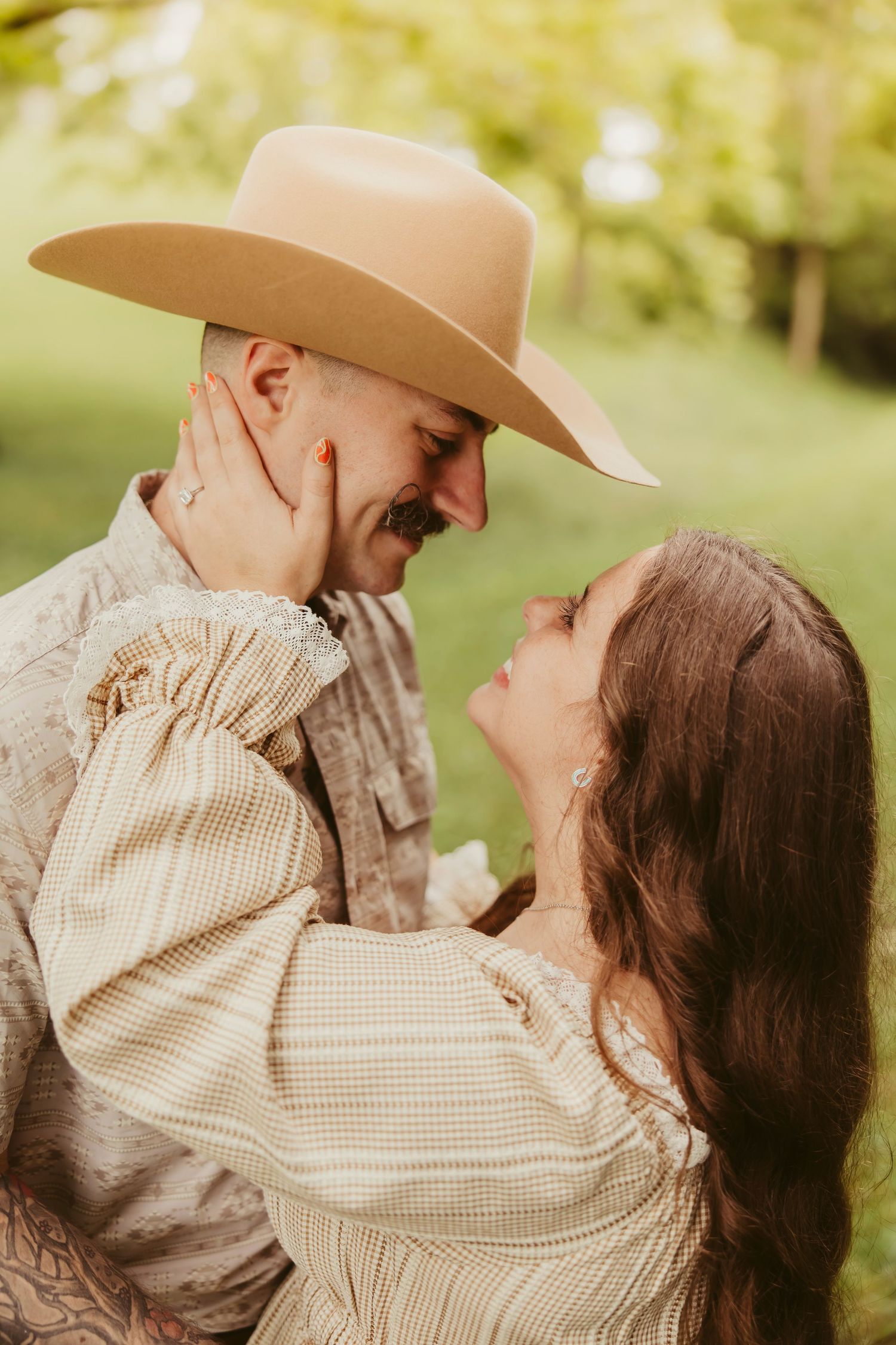 A couple shares a romantic moment outdoors, man in cowboy hat and woman in vintage dress gazing at each other.
