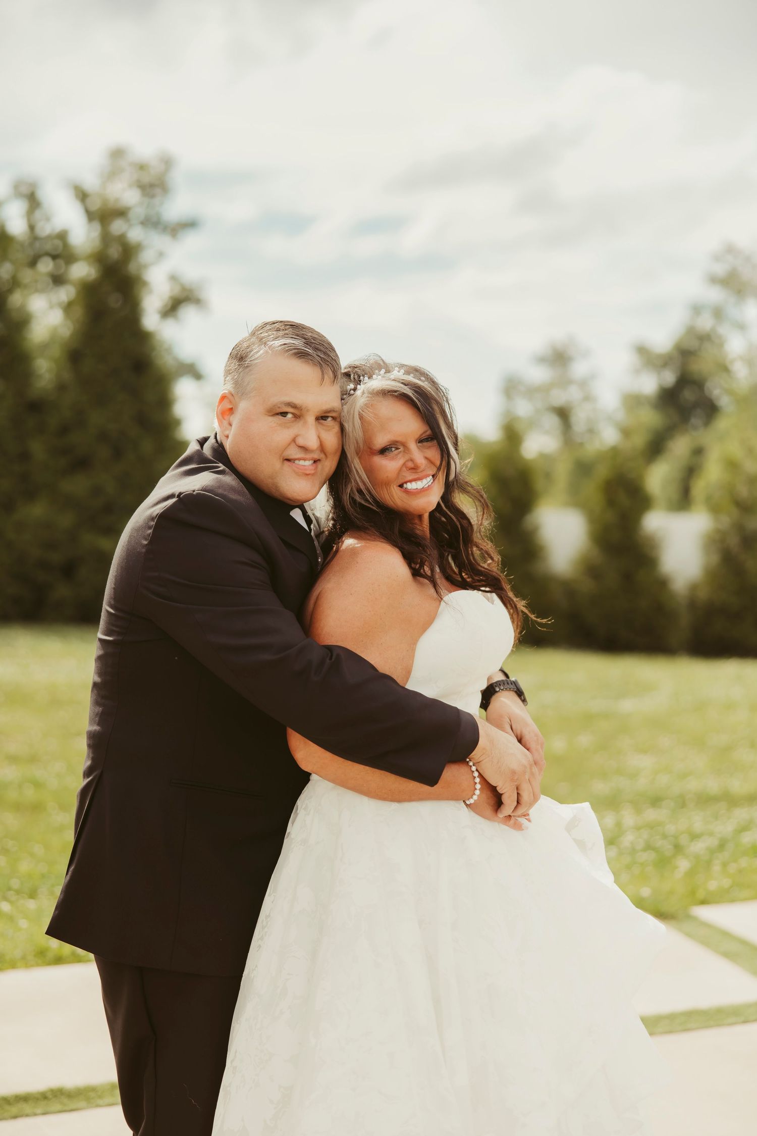 Happy bride and groom embracing outdoors on wedding day, bride in strapless white gown, groom in black suit.
