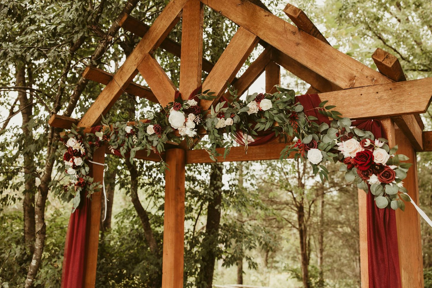 Rustic wooden wedding arch decorated with burgundy and white floral arrangements and red fabric draping outdoors.