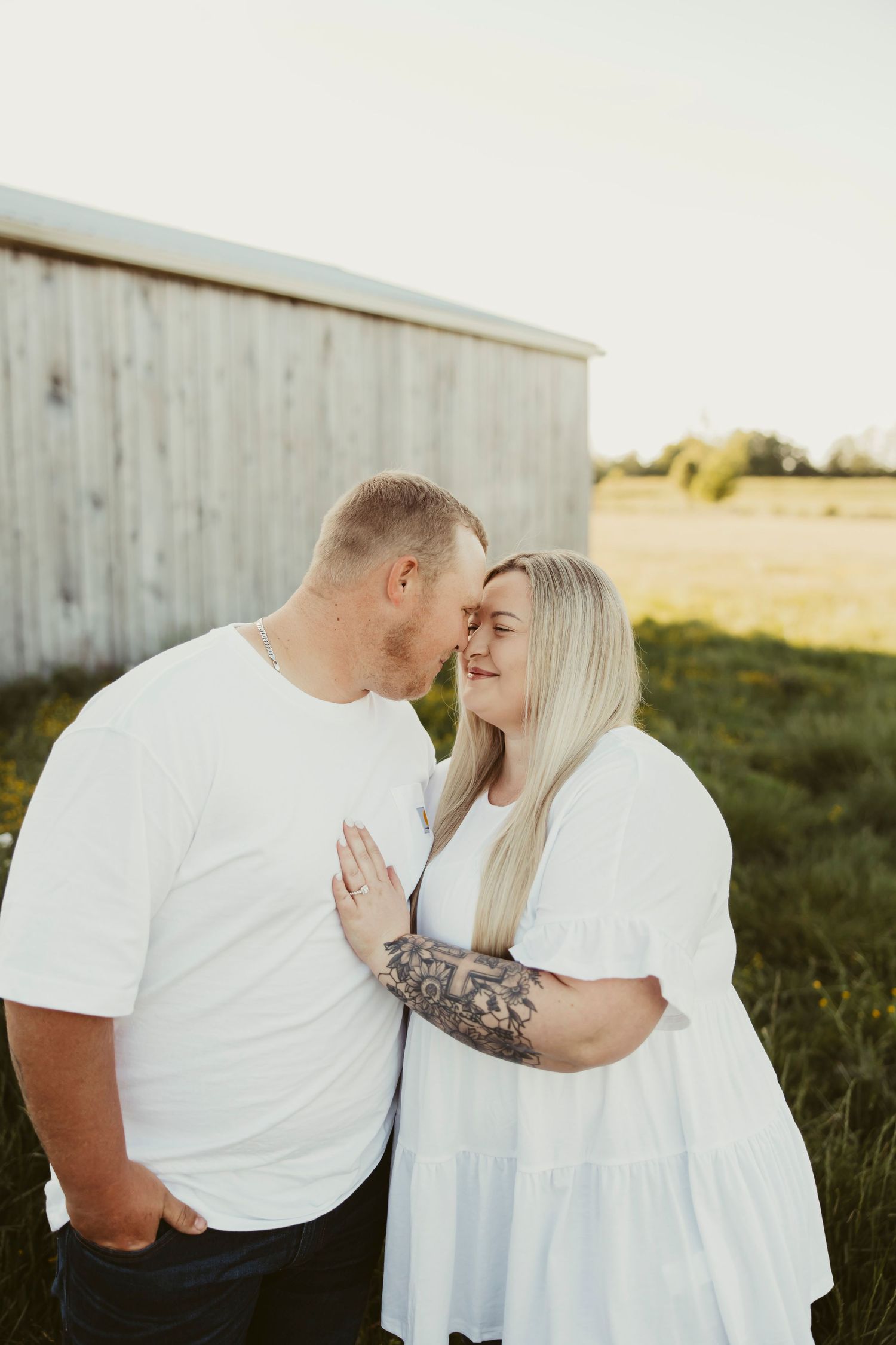 A couple in matching white outfits share a tender moment near a rustic barn in a sunlit field.