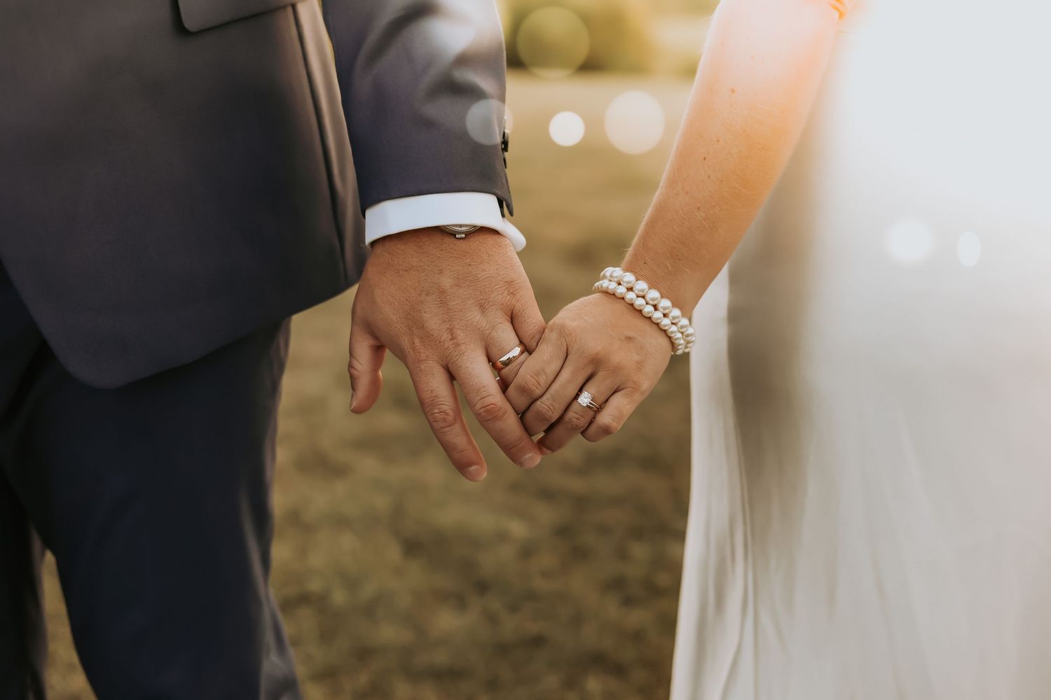 Close-up shot of newlywed couple holding hands showing wedding rings during outdoor ceremony at sunset.
