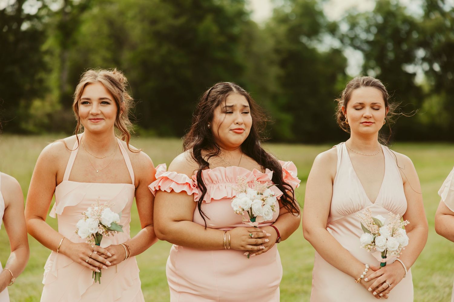Three bridesmaids in blush pink dresses holding white floral bouquets standing outdoors on green grass.