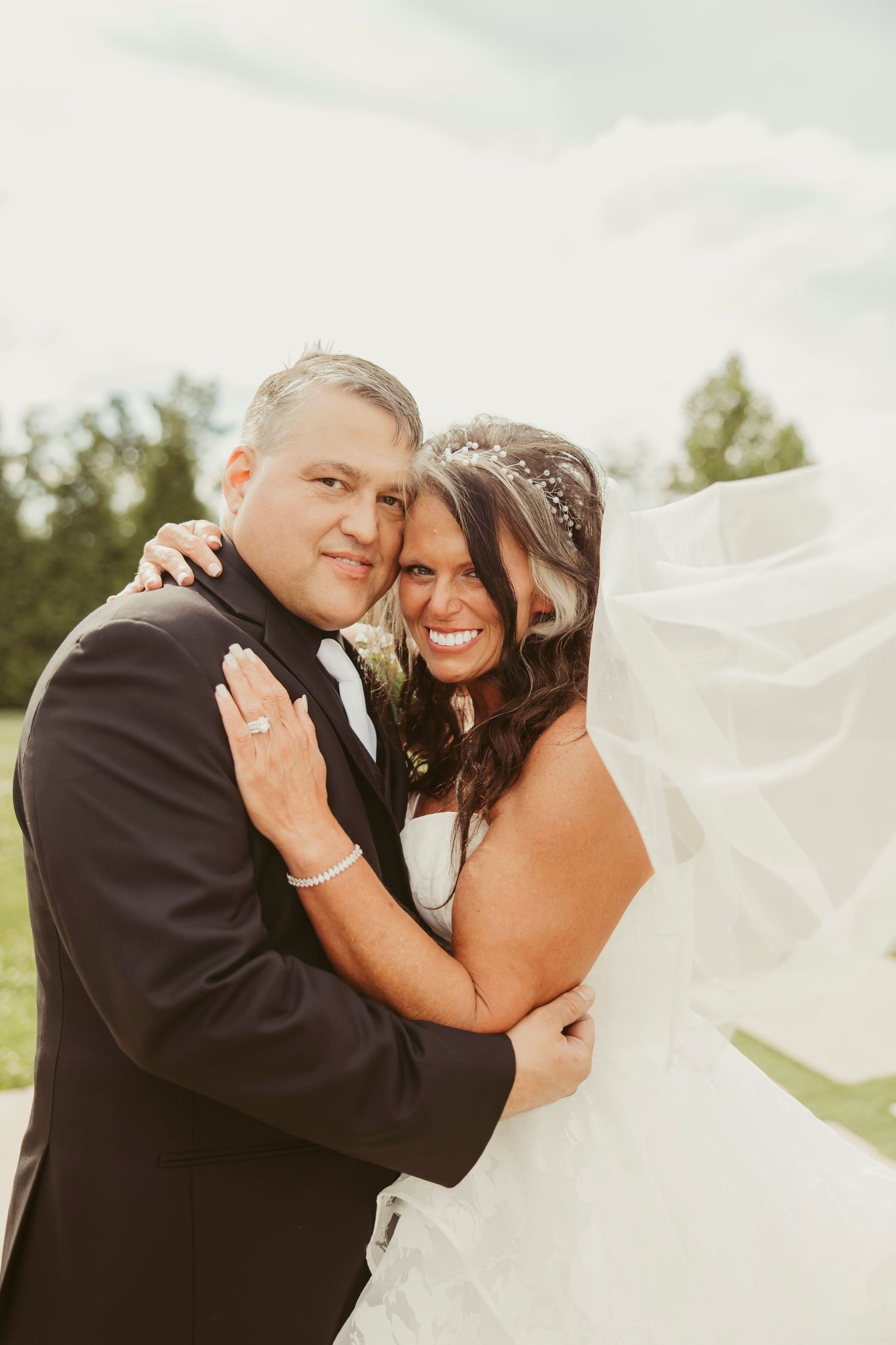 Bride and groom embracing outdoors, her veil flowing in the wind, both smiling joyfully in wedding attire.