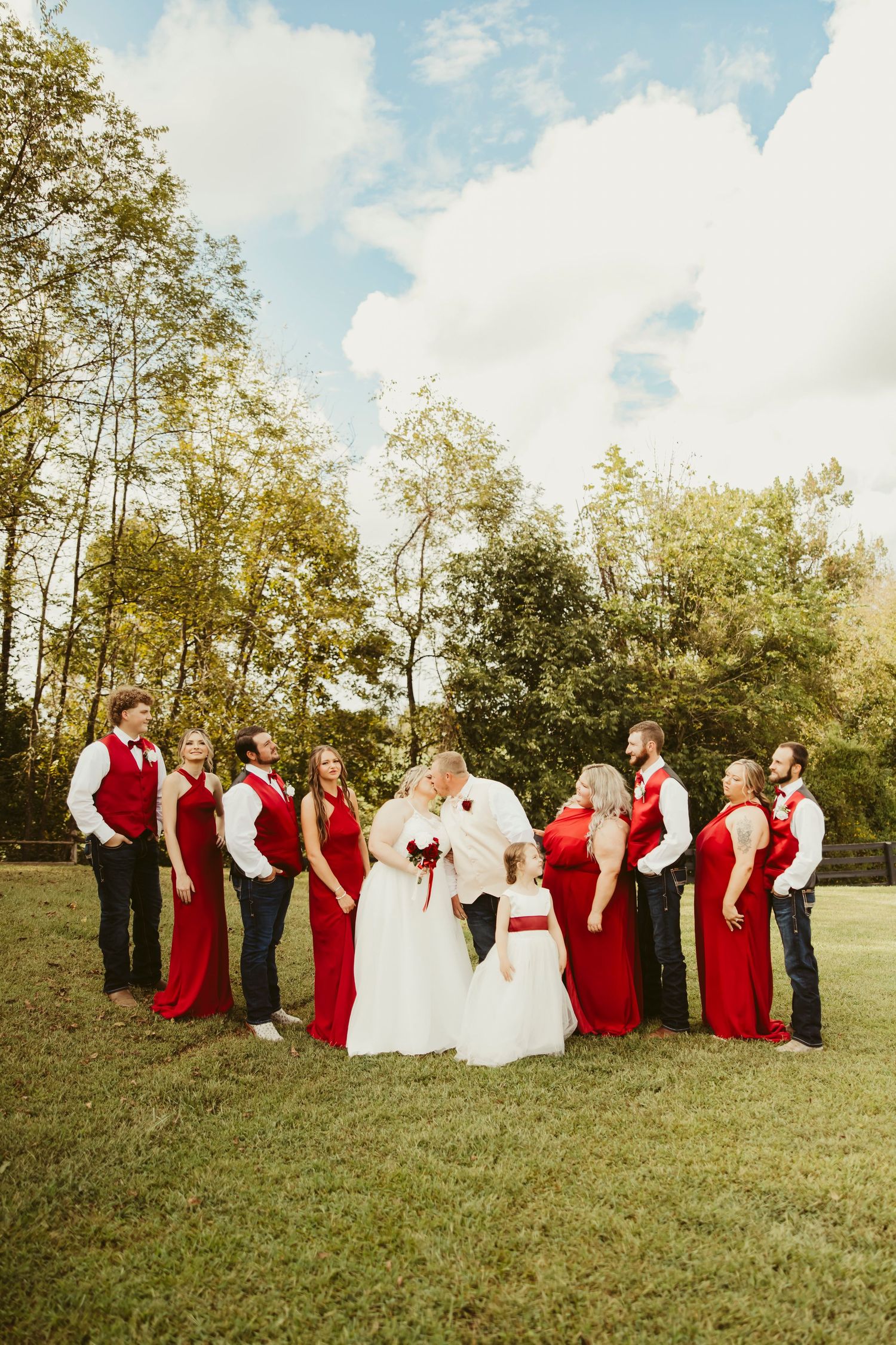 Wedding party poses outdoors with bride and groom kissing, bridesmaids in red gowns and groomsmen in red vests on green lawn.