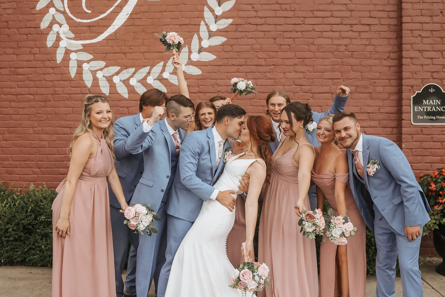 Wedding party in blue suits and pink dresses poses playfully against brick wall.