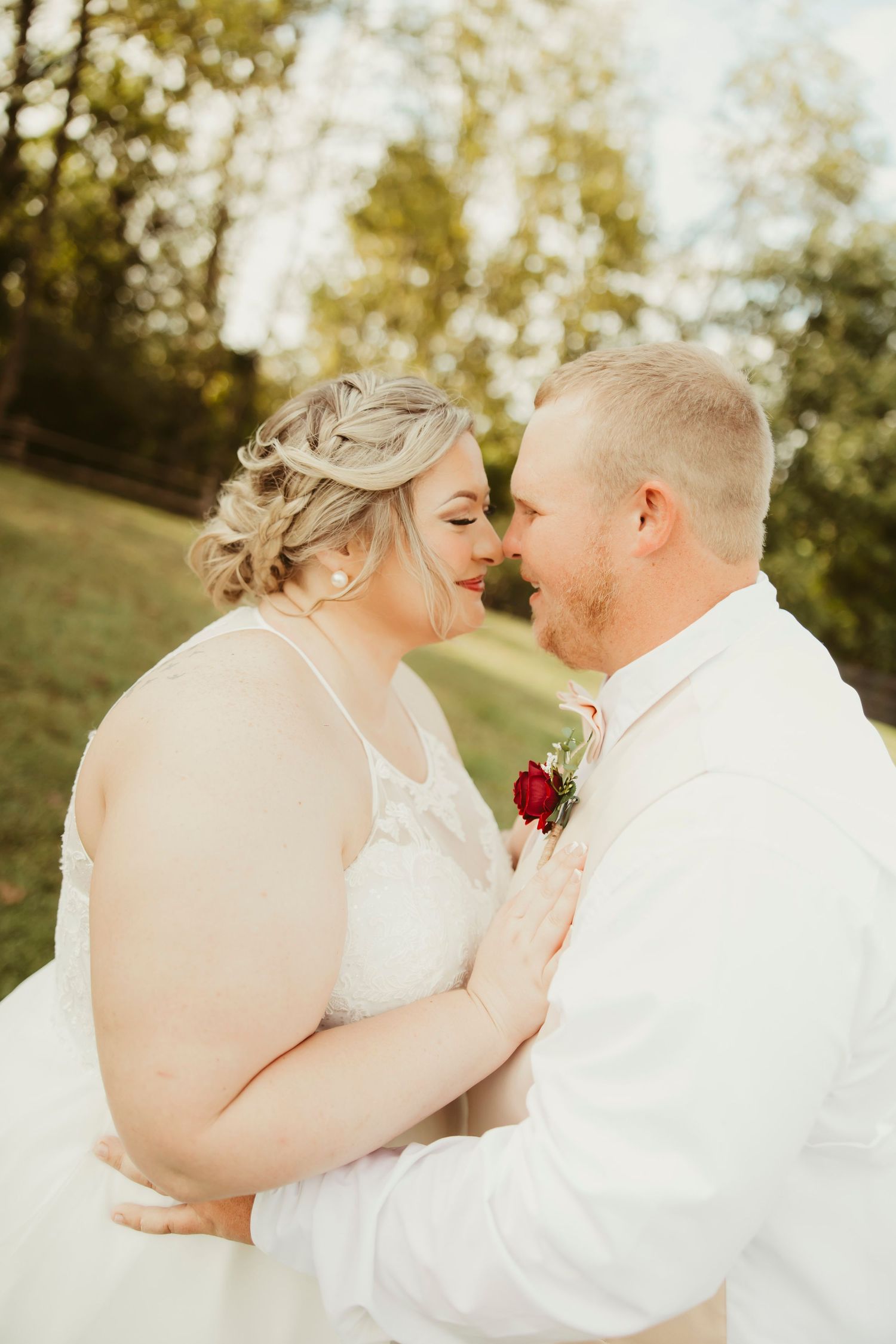 A bride and groom share a romantic moment outdoors, nearly kissing, with the groom wearing a red rose boutonniere.