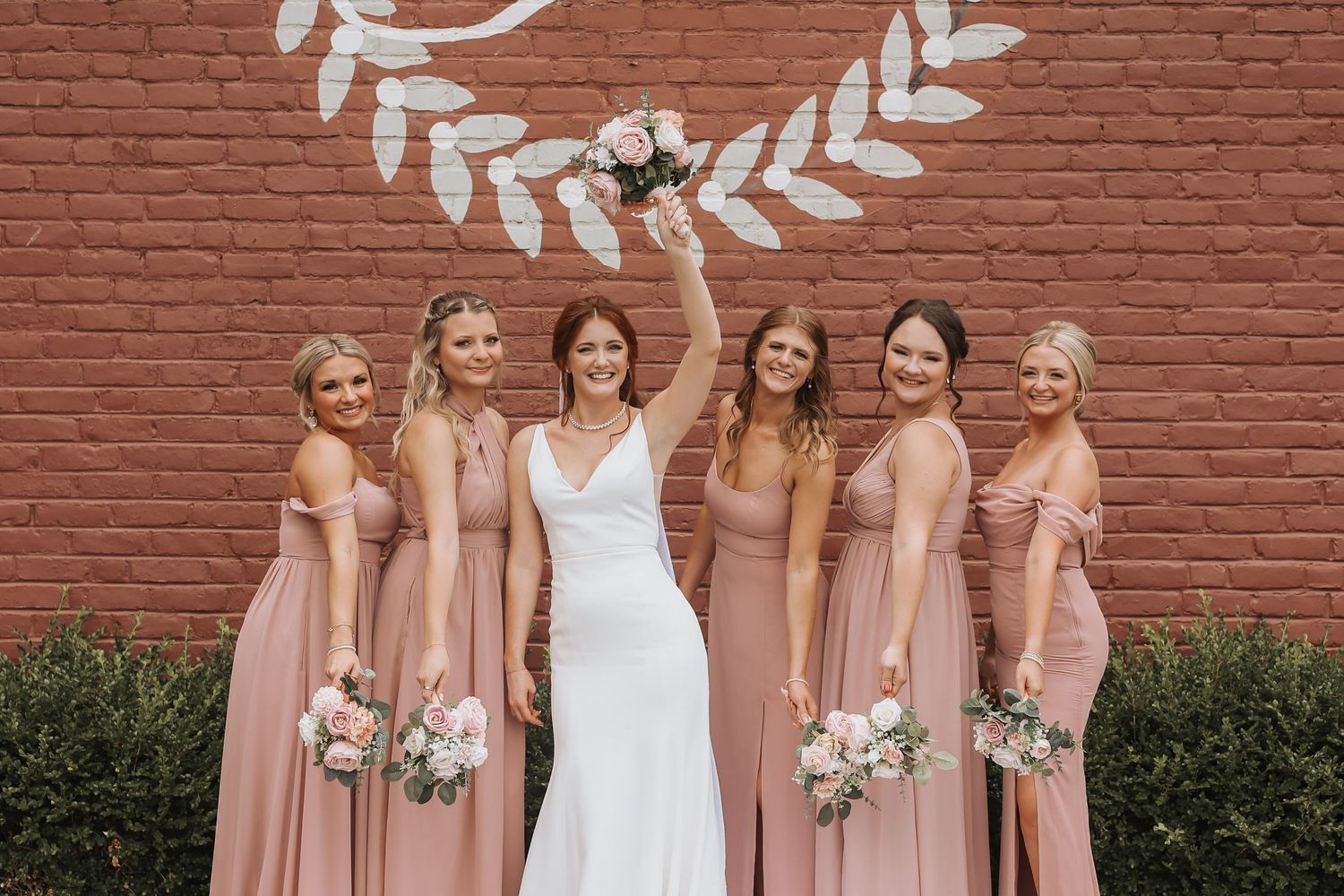 Bridal party wearing matching pink gowns pose together against rustic brick backdrop.