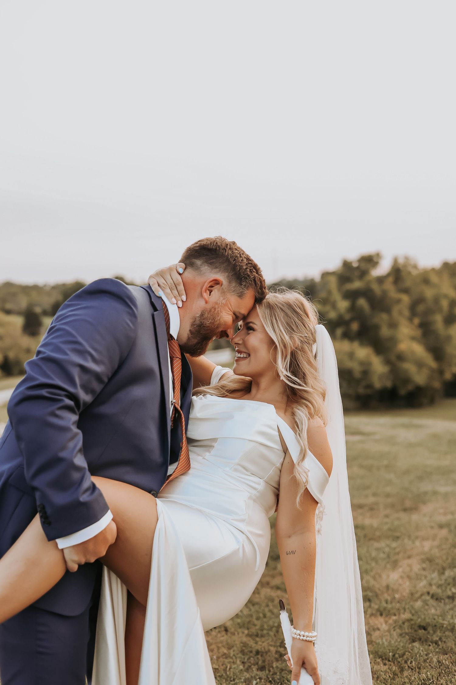 A romantic wedding couple embraces in a scenic outdoor field with a flowing white dress and navy suit at sunset.