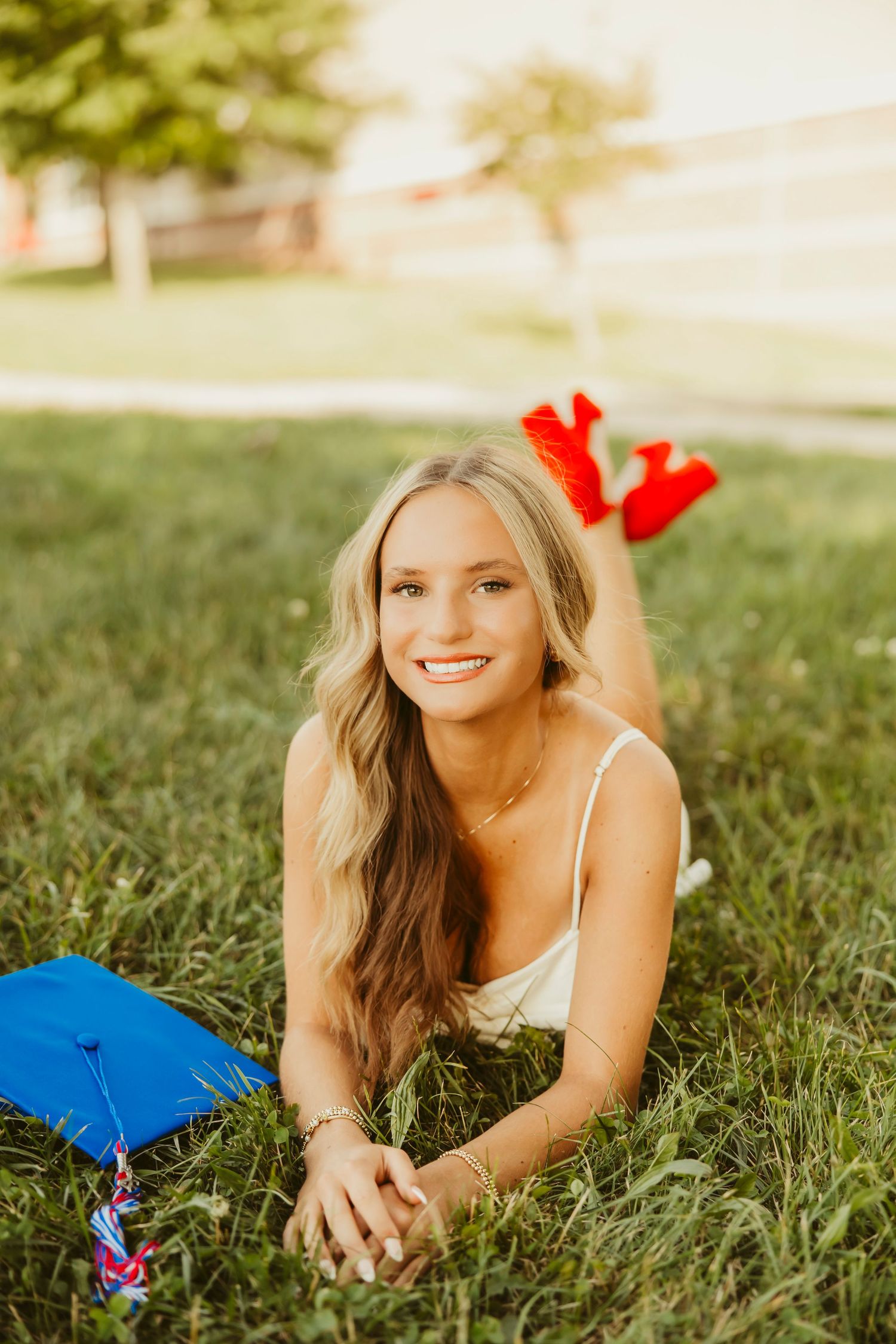 Smiling blonde graduate lying on green grass with blue graduation cap and red tassels in sunny outdoor setting.