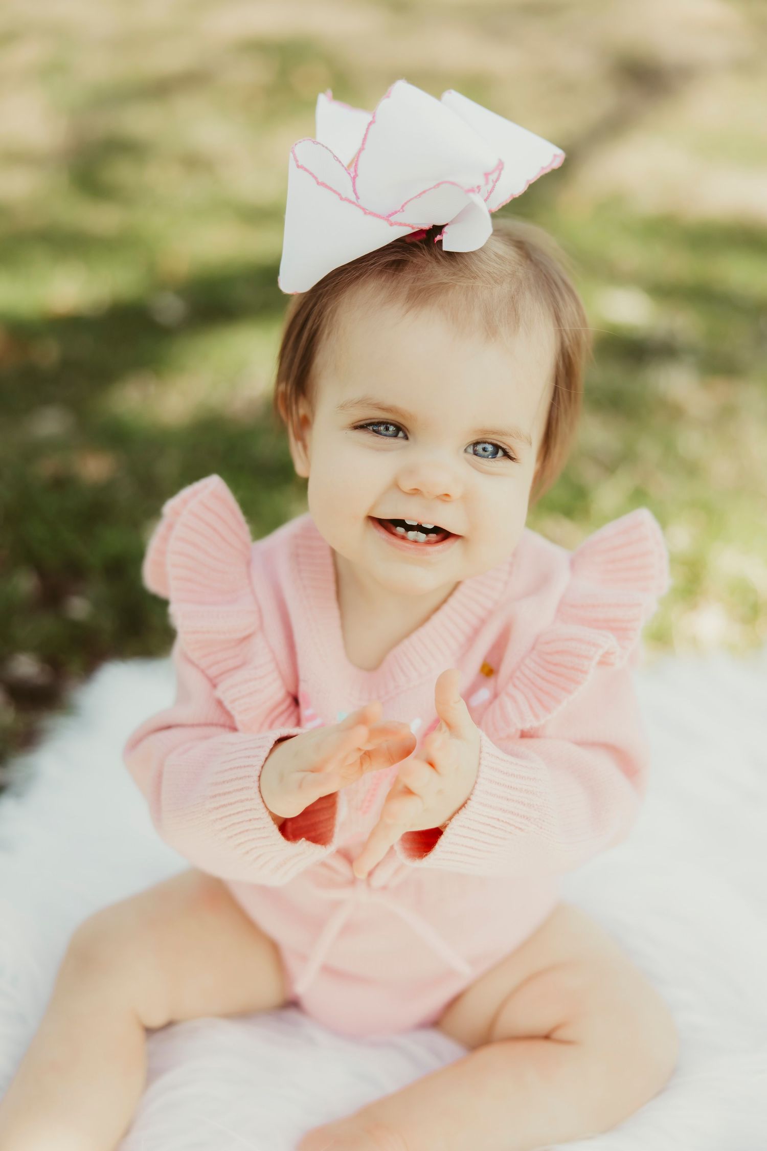 Smiling baby girl in pink ruffle romper with white bow headband sitting outdoors on white blanket holding apple slice.