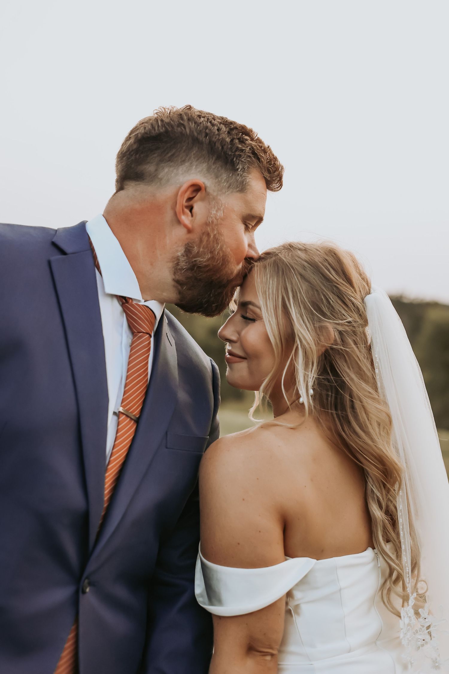 A tender wedding moment captured outdoors with a couple in navy suit and off-shoulder white gown with veil.