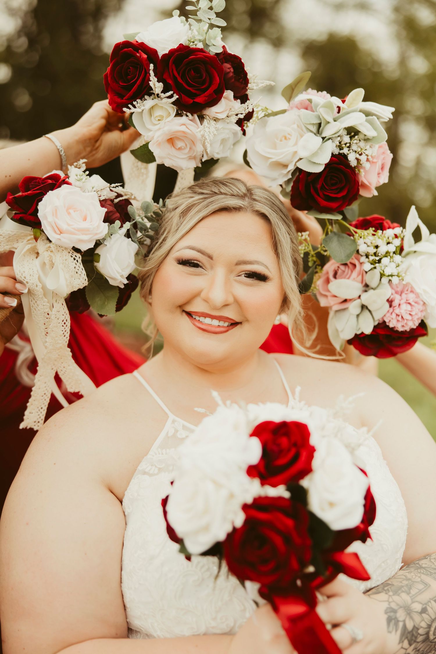 Smiling bride holding red and white rose bouquet surrounded by bridesmaids in red dresses lifting floral arrangements.