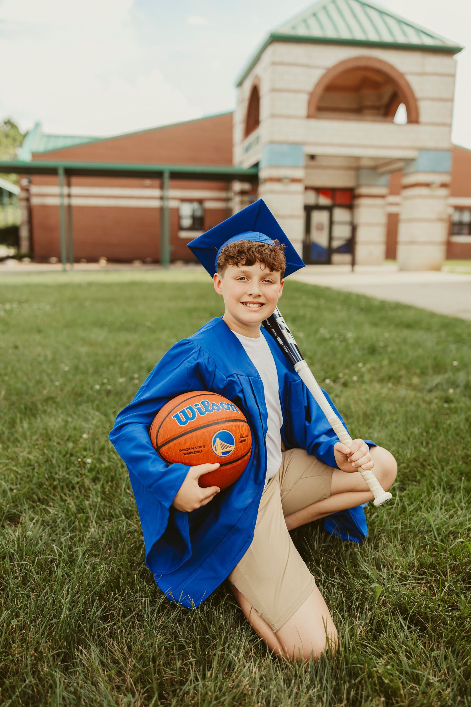 A smiling boy in a blue graduation cap and gown holds a Wilson basketball and diploma while kneeling on grass.