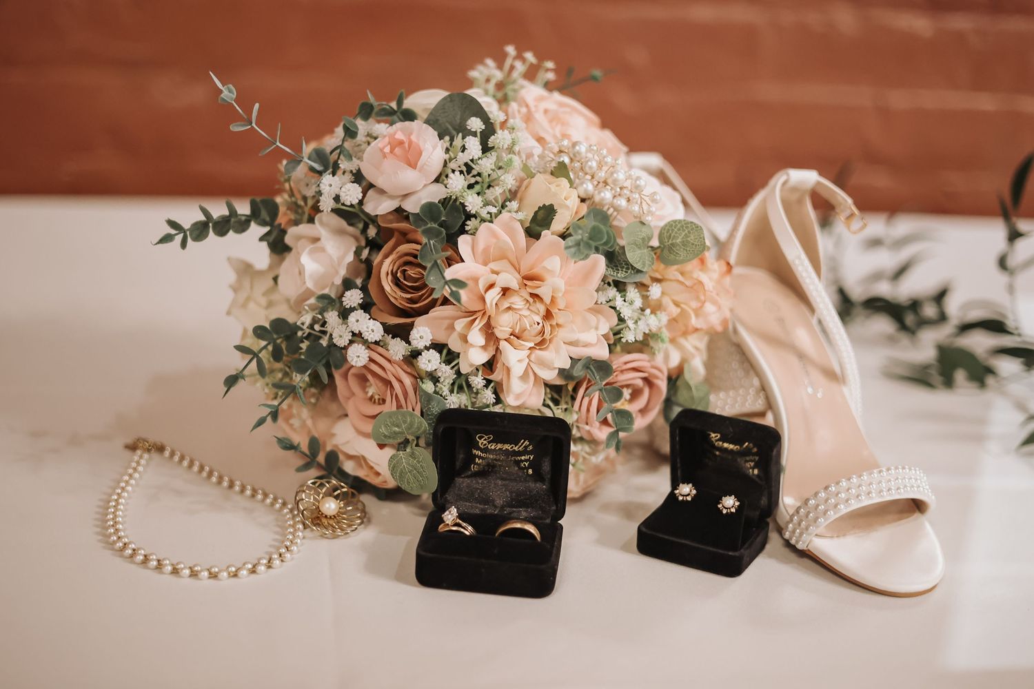 Wedding details featuring a blush rose bouquet, pearl necklace, and ring boxes on white surface.