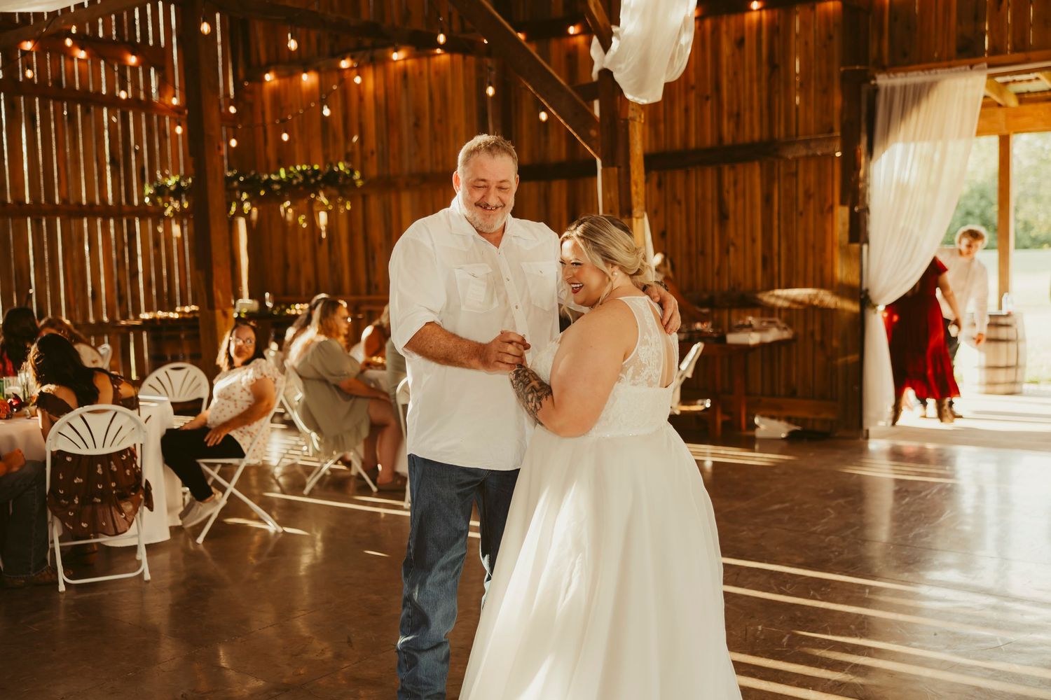 Bride and groom share first dance in rustic barn wedding venue with string lights and guests seated at tables.