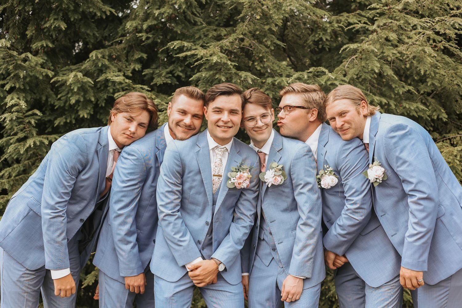 Groomsmen in light blue suits pose together in front of evergreen trees.