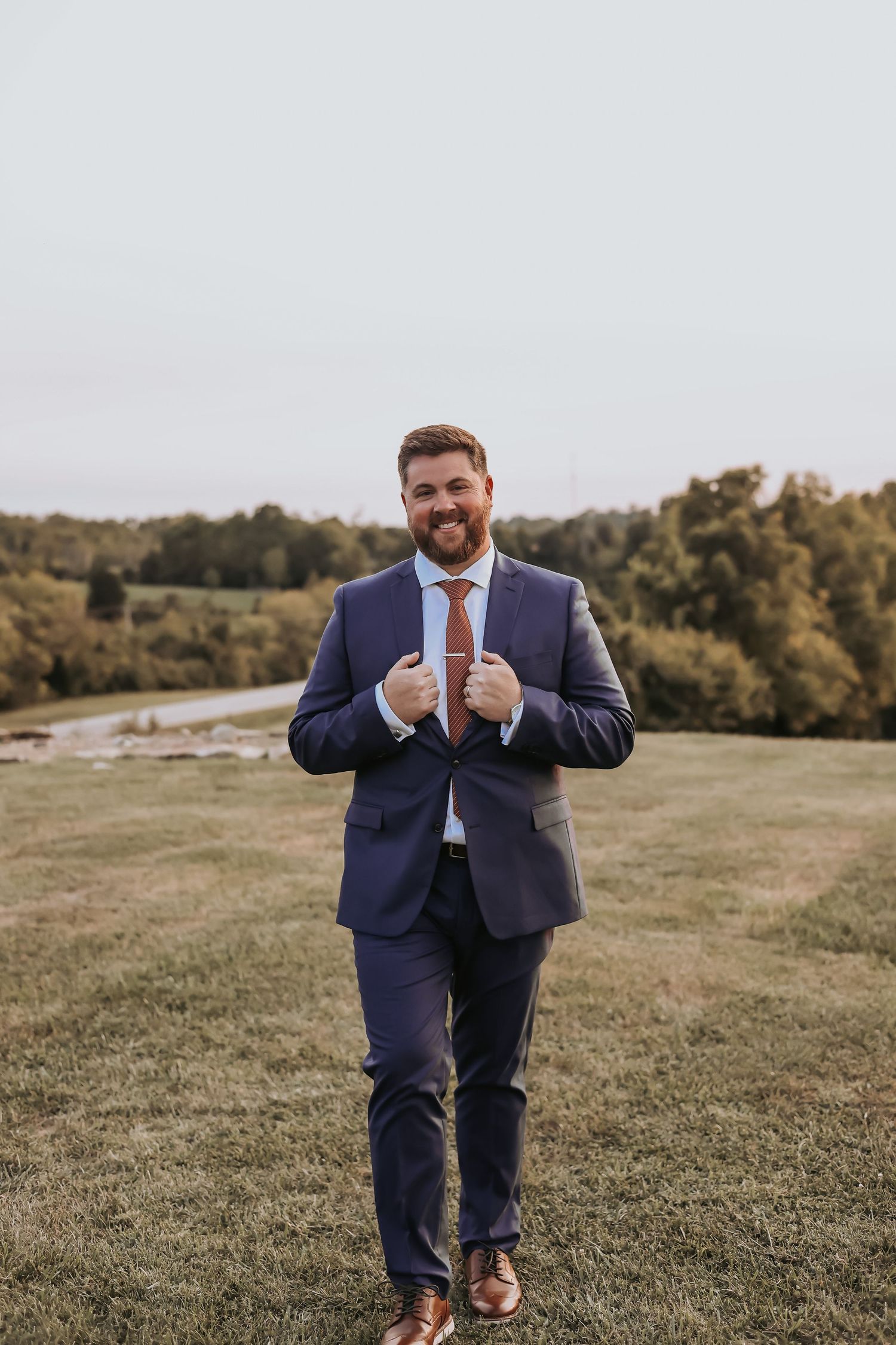A groom in a navy blue suit and orange tie adjusts his jacket while standing in a grassy outdoor setting.