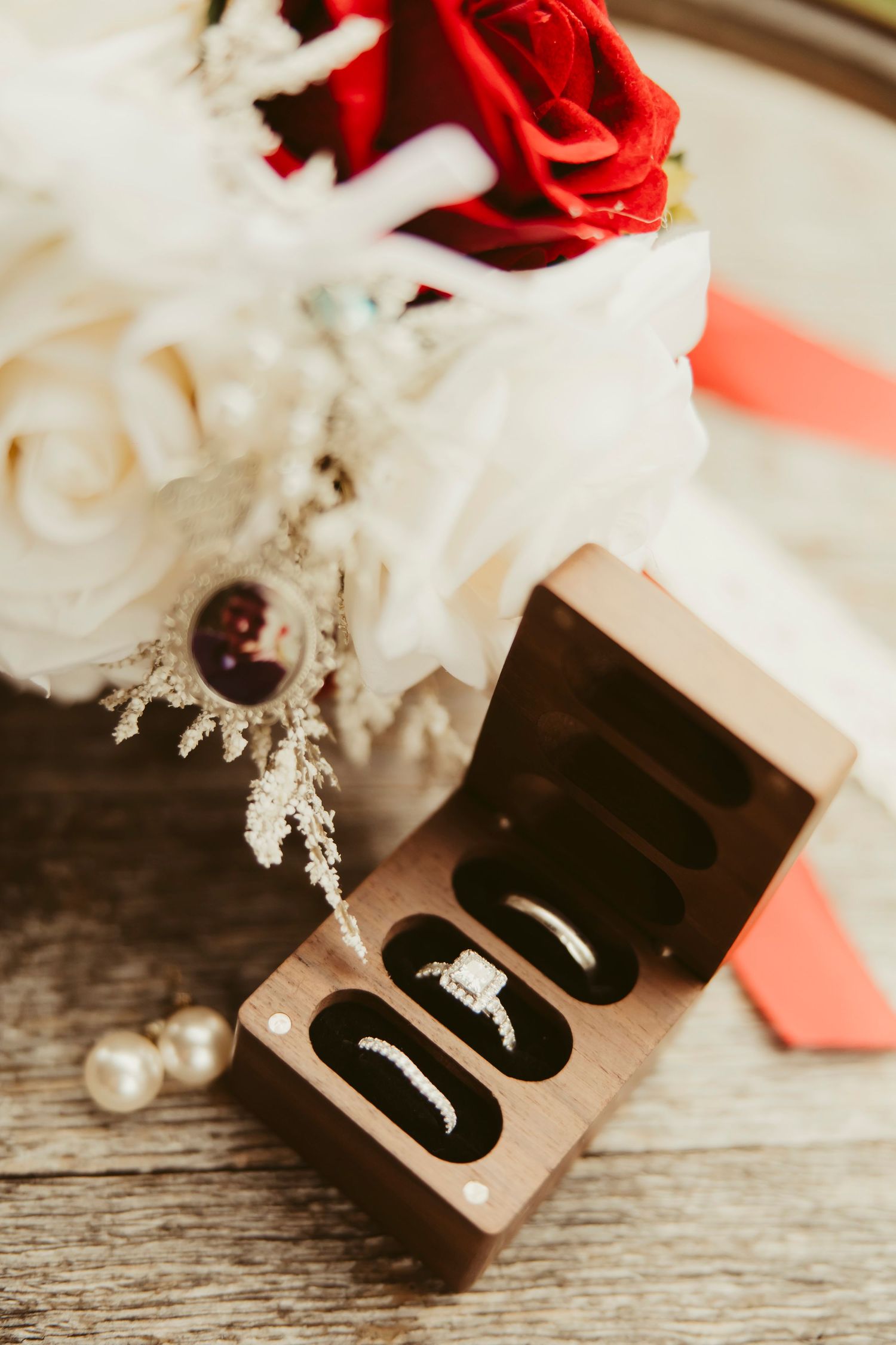 Wedding rings displayed in a wooden box beside a red and white rose bridal bouquet and pearl earrings.