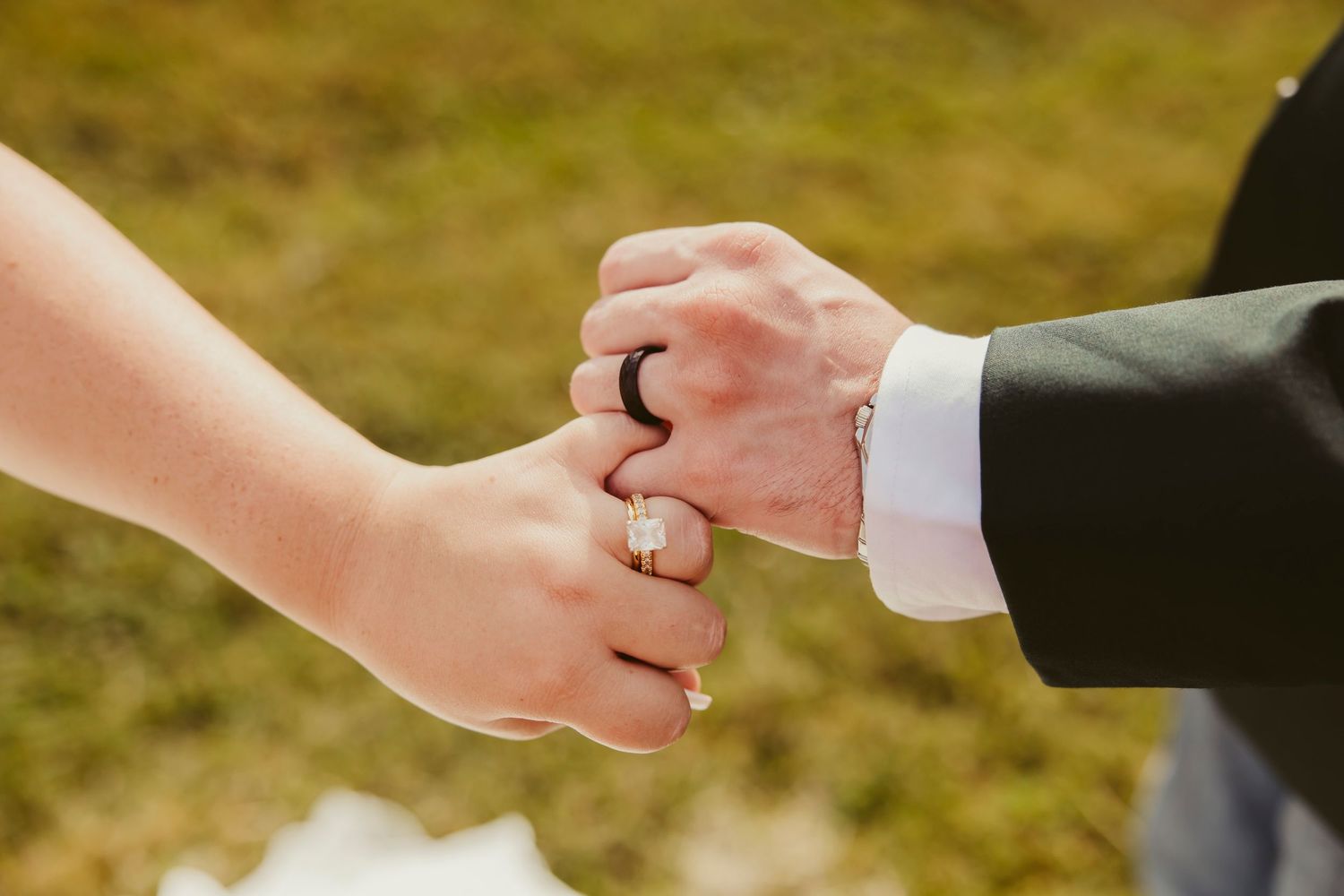 Close-up of a couple holding hands showing wedding rings, bride wearing diamond ring, groom wearing black band, outdoor ceremony.