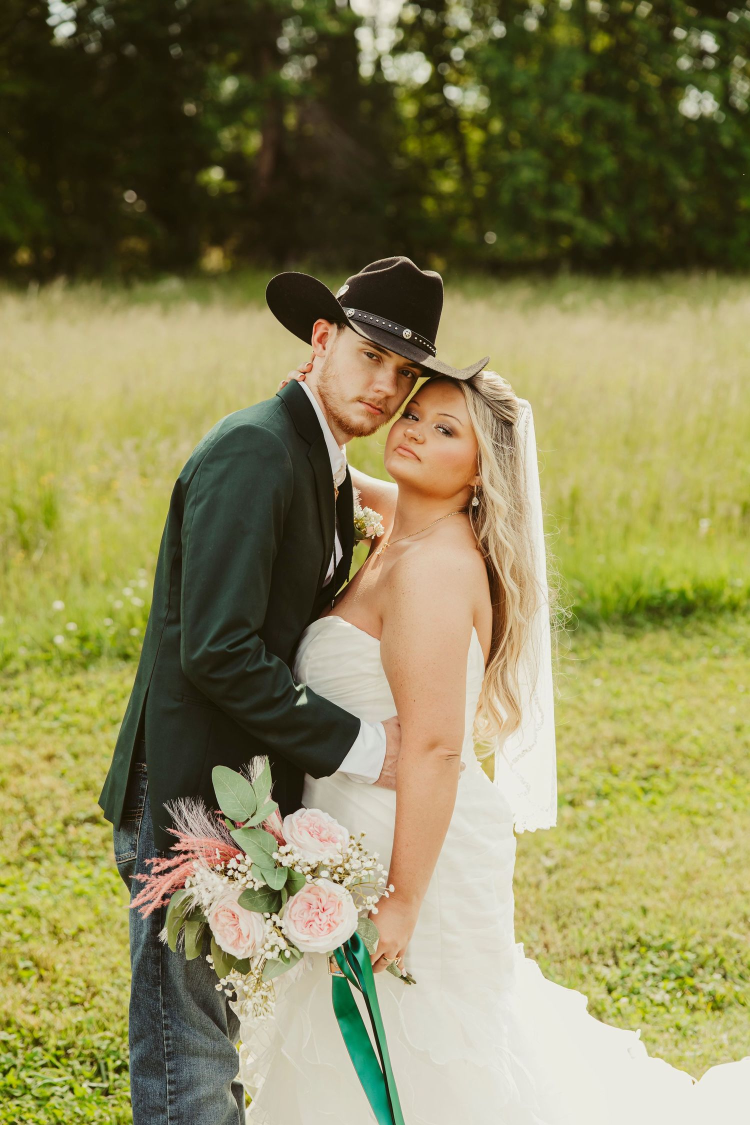 Bride and groom pose in field wearing matching black cowboy hats, bride holding pink floral bouquet.