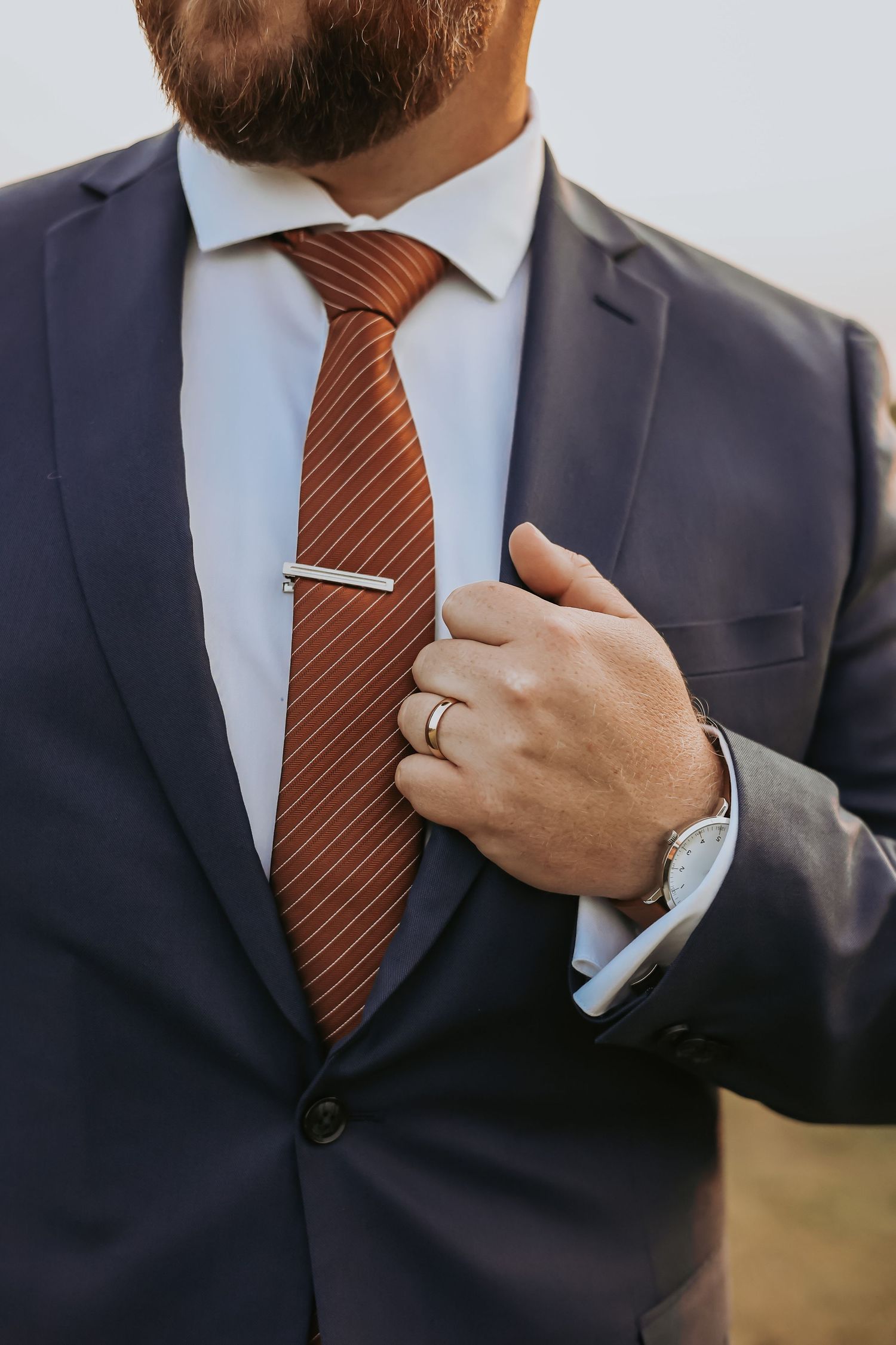 Close-up of groom's navy suit with orange striped tie and silver tie clip.