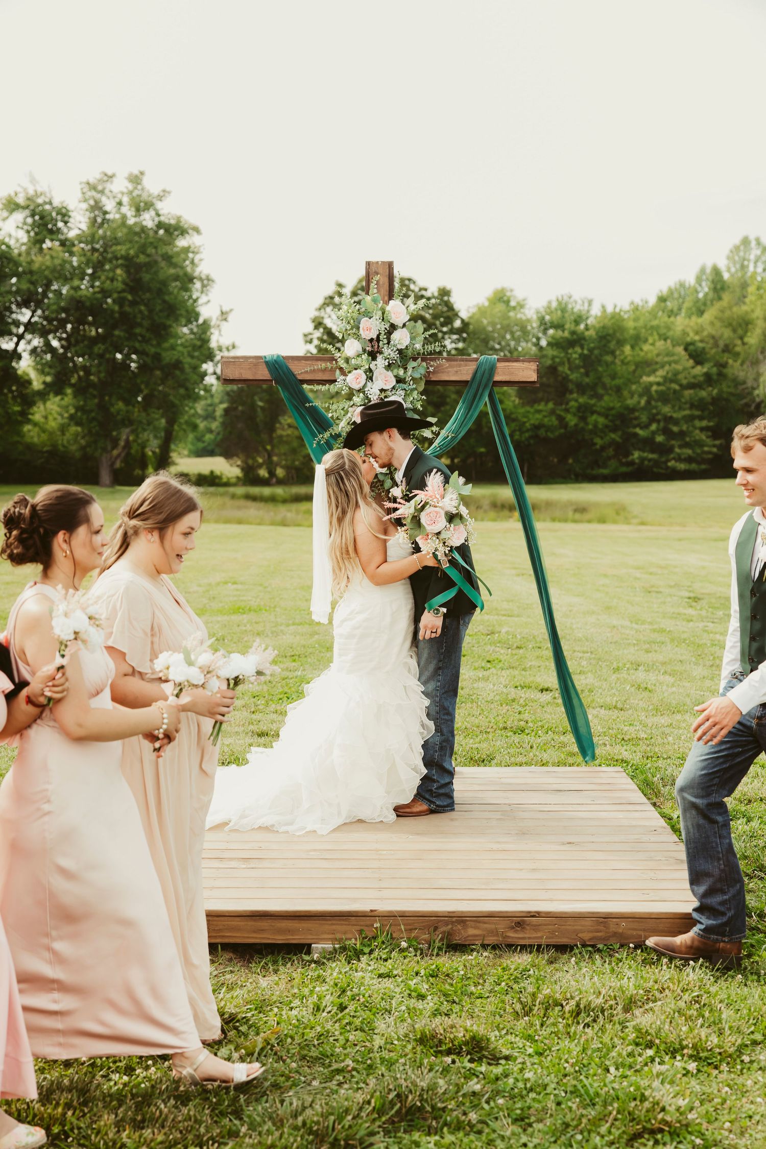Bride and groom share first kiss at outdoor rustic wedding ceremony with teal arch, pink florals, and bridesmaids in blush gowns.