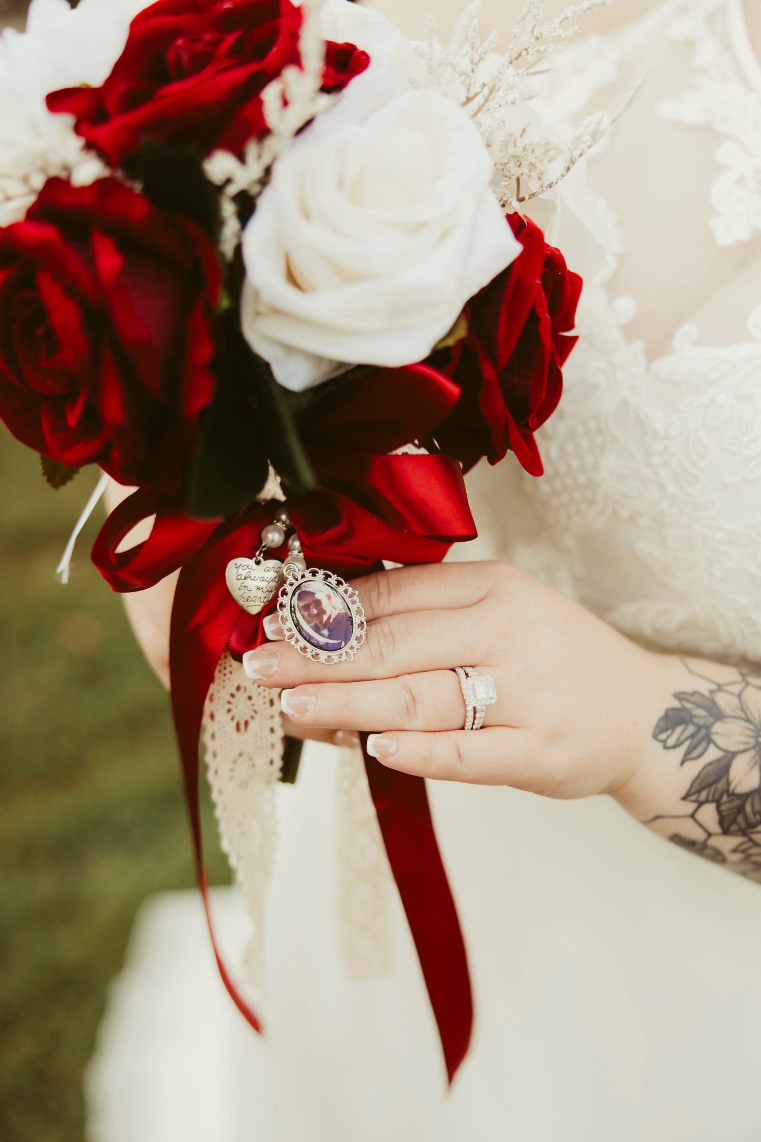 Bride holds red and white rose bouquet with memorial locket charms, wearing wedding rings and lace gown.