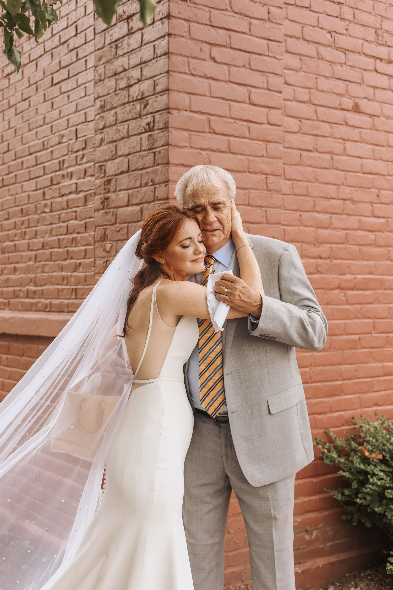 Tender embrace between bride and elderly gentleman against terracotta brick wall.