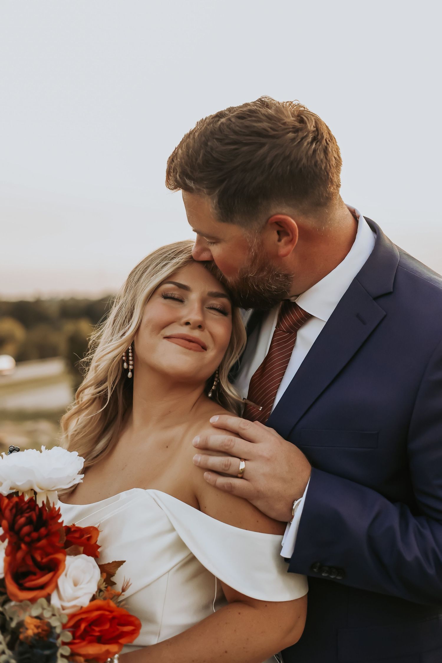 Intimate wedding portrait shows tender moment between couple with red and white floral bouquet.
