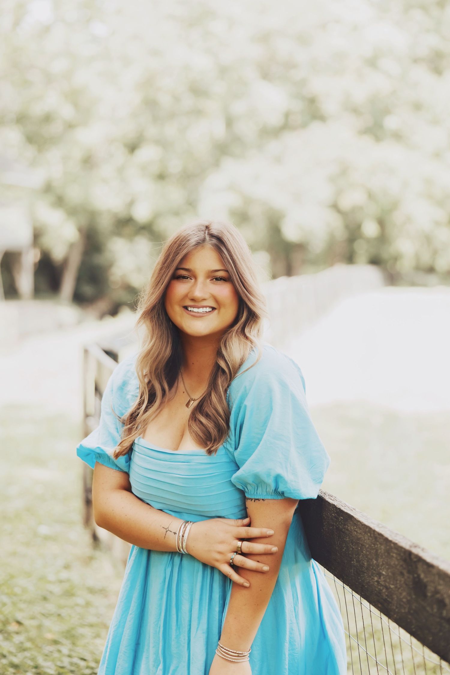 Young woman in a light blue puff-sleeve dress smiling outdoors near a wooden fence with green trees.
