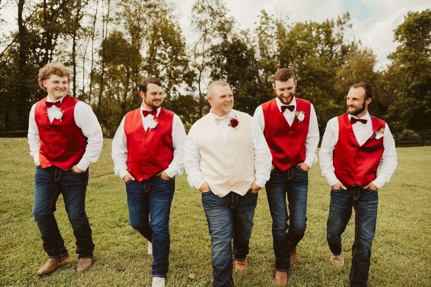 Groom in white shirt walks with four groomsmen wearing red vests and bow ties on a green lawn.